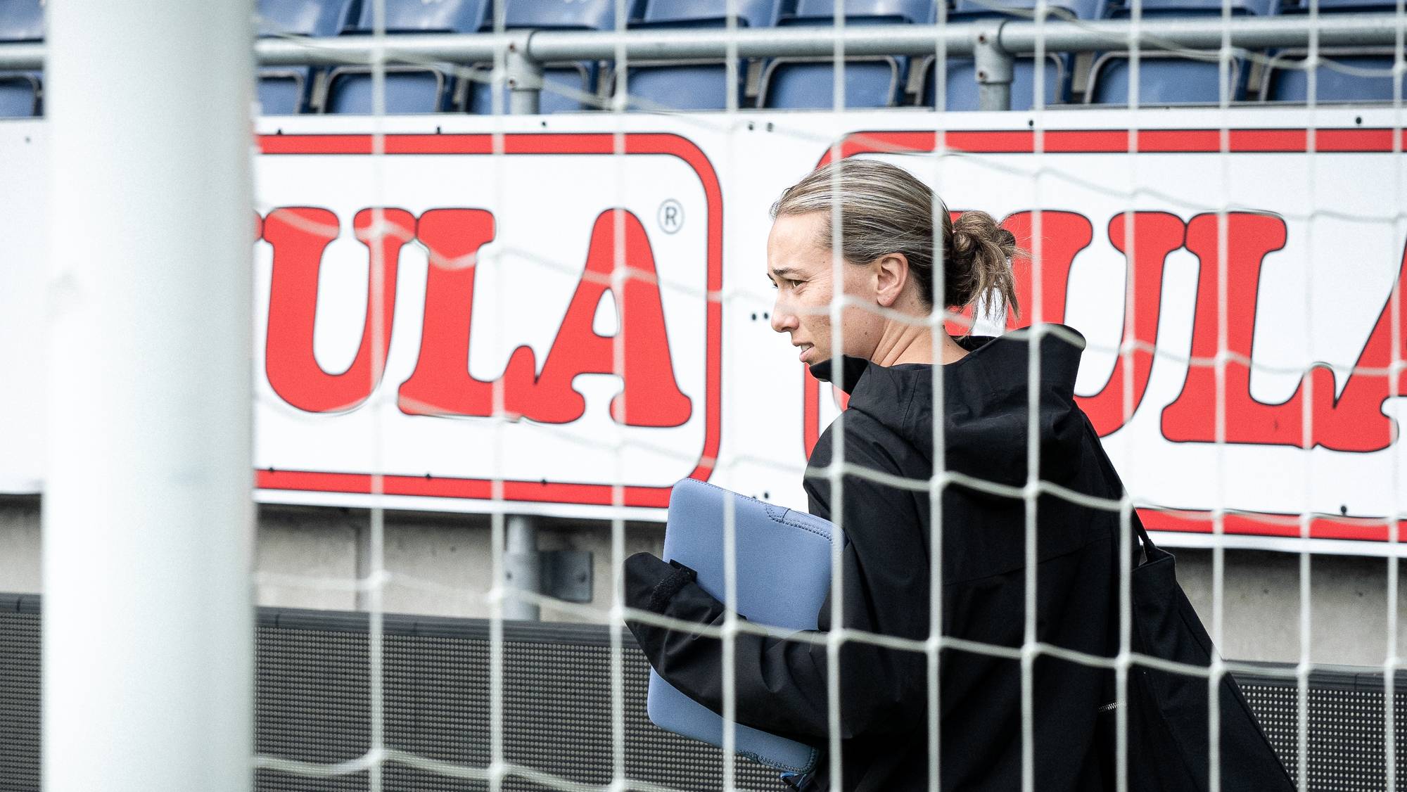 Katarina Dybvik Sunde i sort jakke med mappe går langs en fotballbane sett gjennom et målnett. 