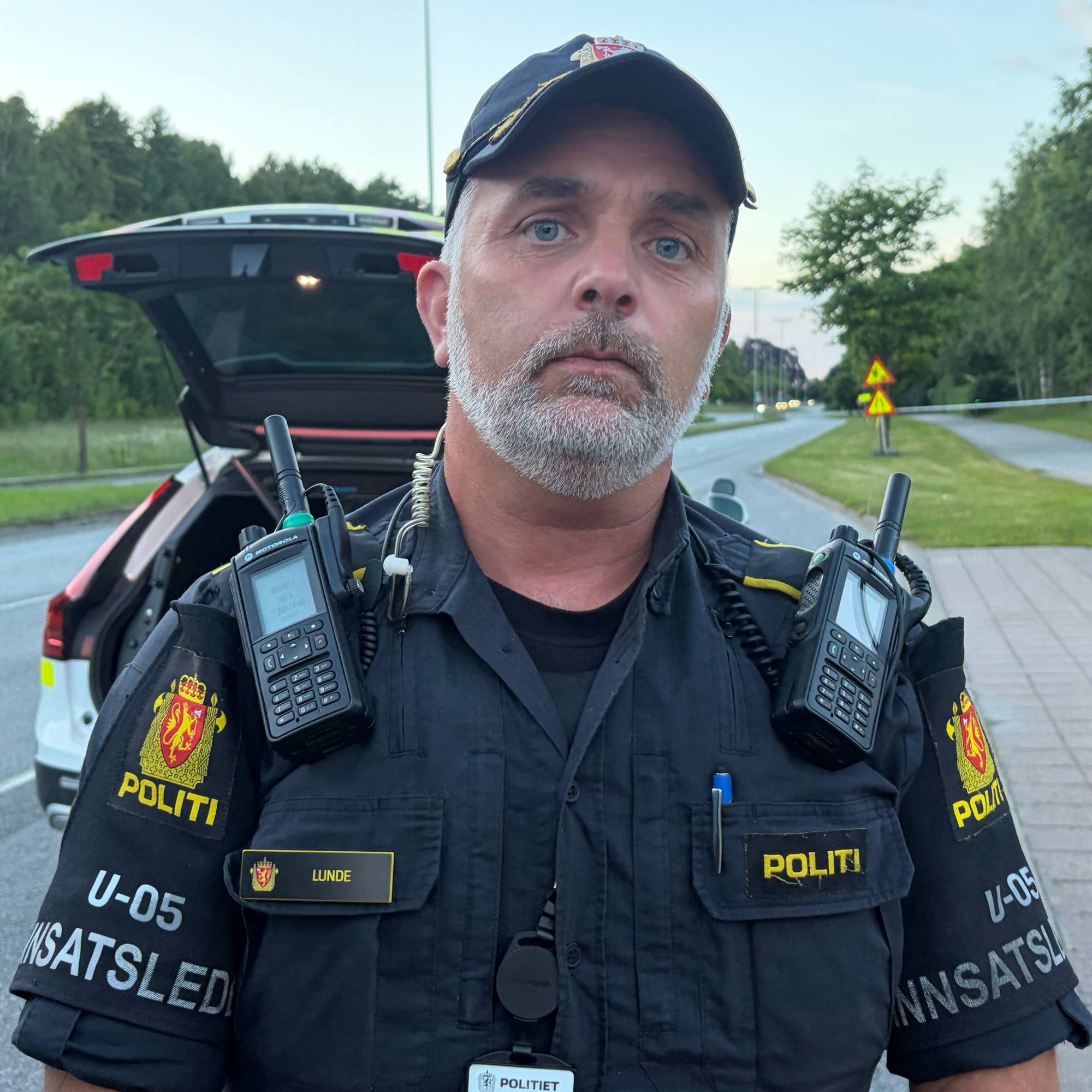 En politi-innsatsleder står ved en vei med en politibil i bakgrunnen. (Bildebeskrivelsen er laget av en KI-tjeneste)