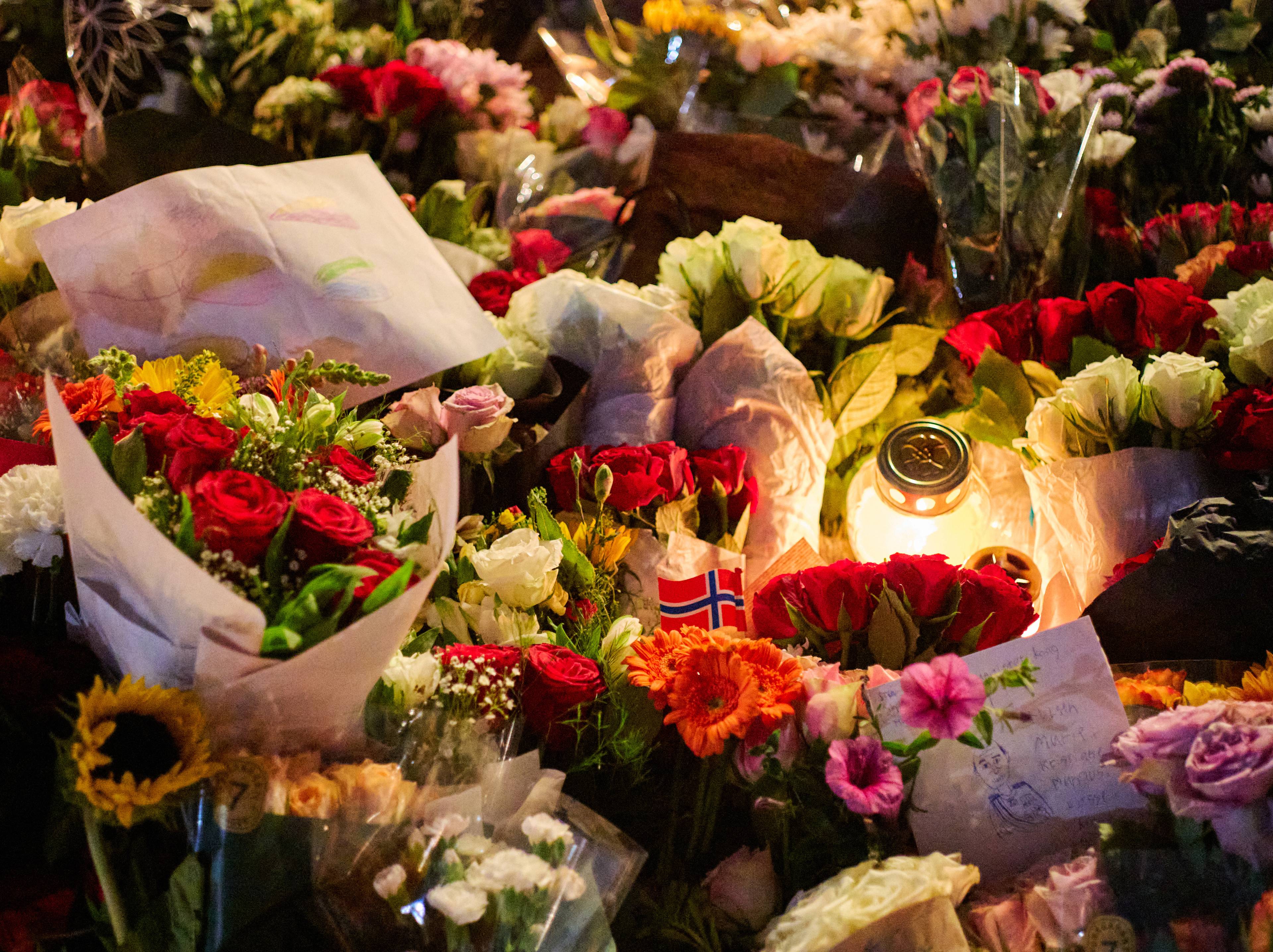 Mange blomsterbuketter og et tent lys er plassert som en minneplass. Et lite norsk flagg er synlig.
