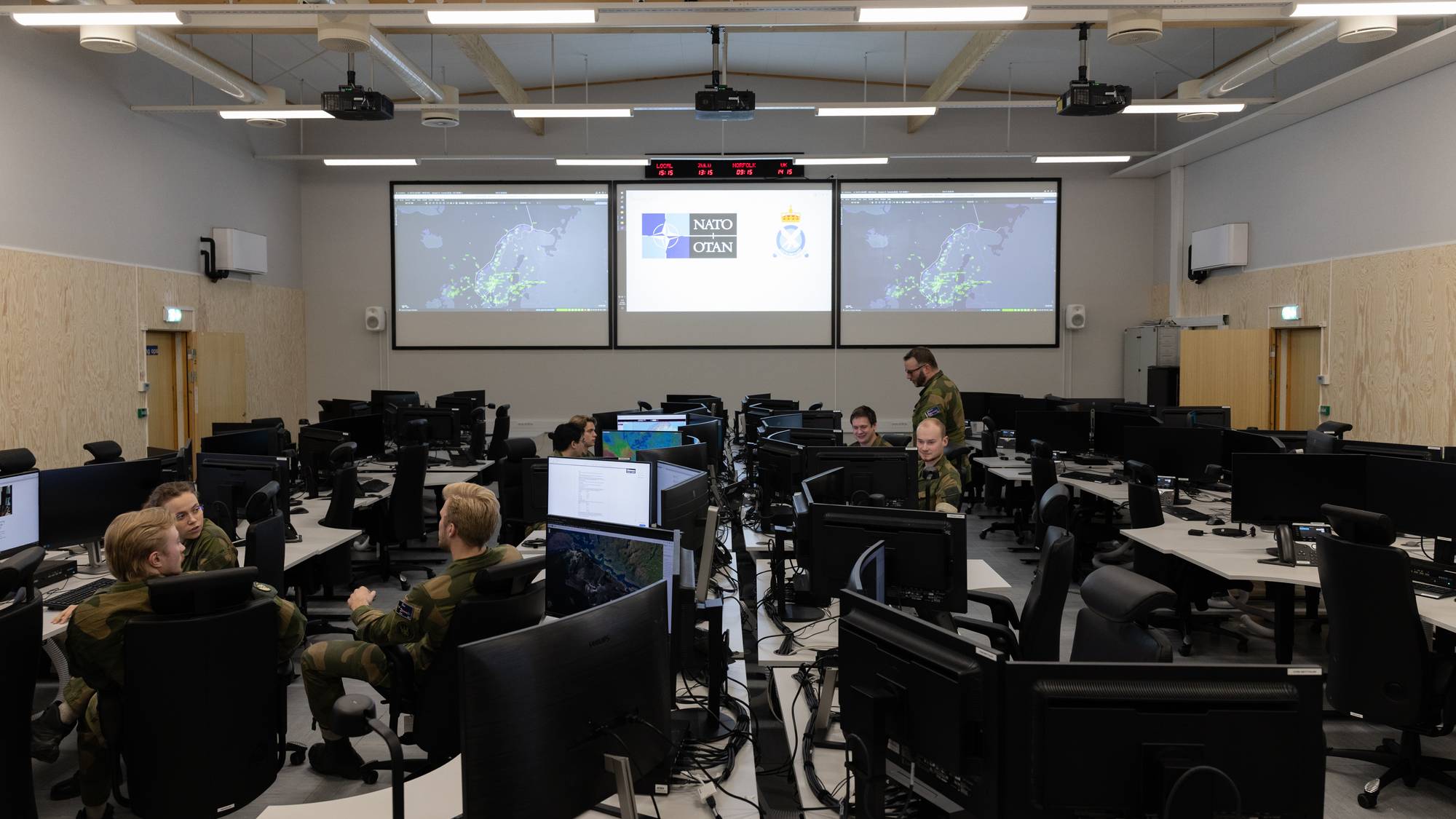 Flere soldater sitter ved datamaskiner i et rom med store skjermer. På skjermene vises kartdata. Et NATO-flagg og det norske kongeflagget er synlige. Rommet har moderne innredning med trevegger og himling. (Bildebeskrivelsen er laget av en KI-tjeneste)