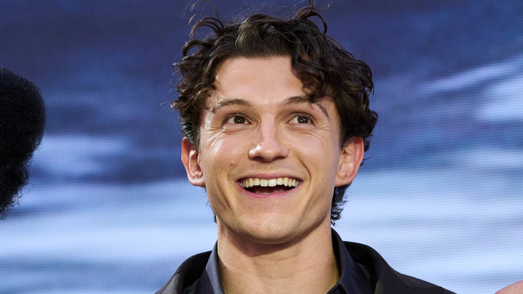 Skuespiller Tom Holland smiler mot kameraet foran en blå bakgrunn. (Bildebeskrivelsen er laget av en KI-tjeneste)