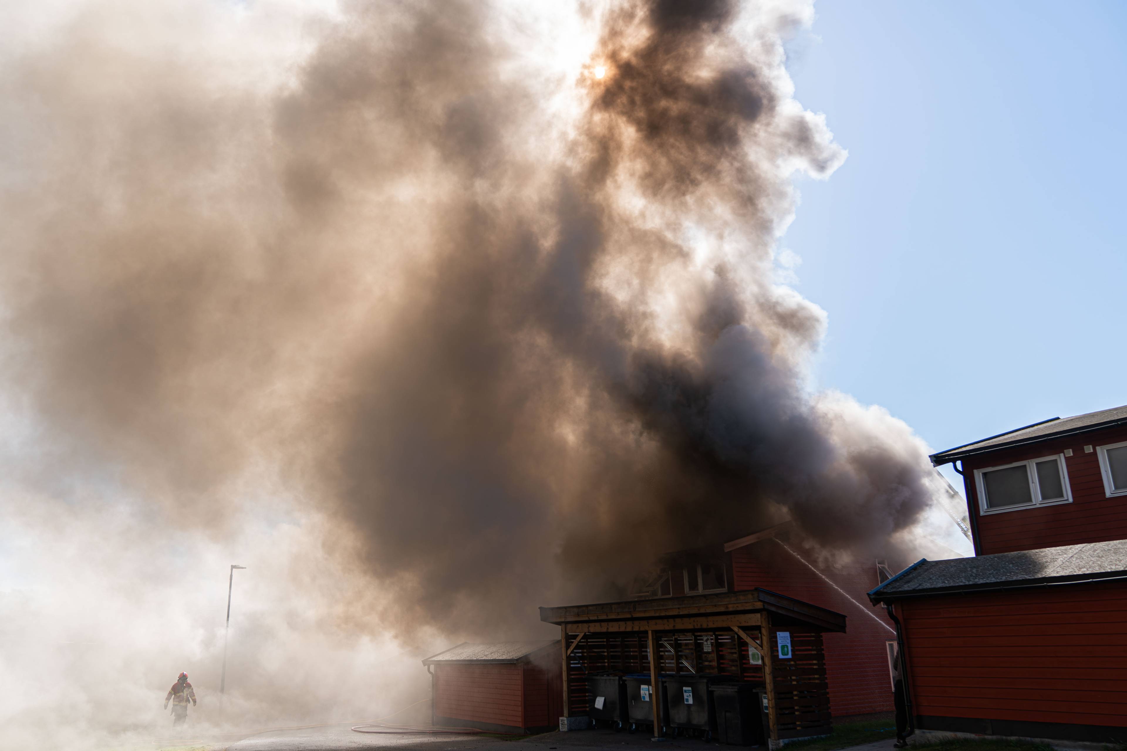 Brannmann i fullt verneutstyr jobber ved en brennende bygning. Det stiger store mengder mørk røyk opp mot en skyfri himmel. Flere røde trebygninger er synlige i bildet. (Bildebeskrivelsen er laget av en KI-tjeneste)