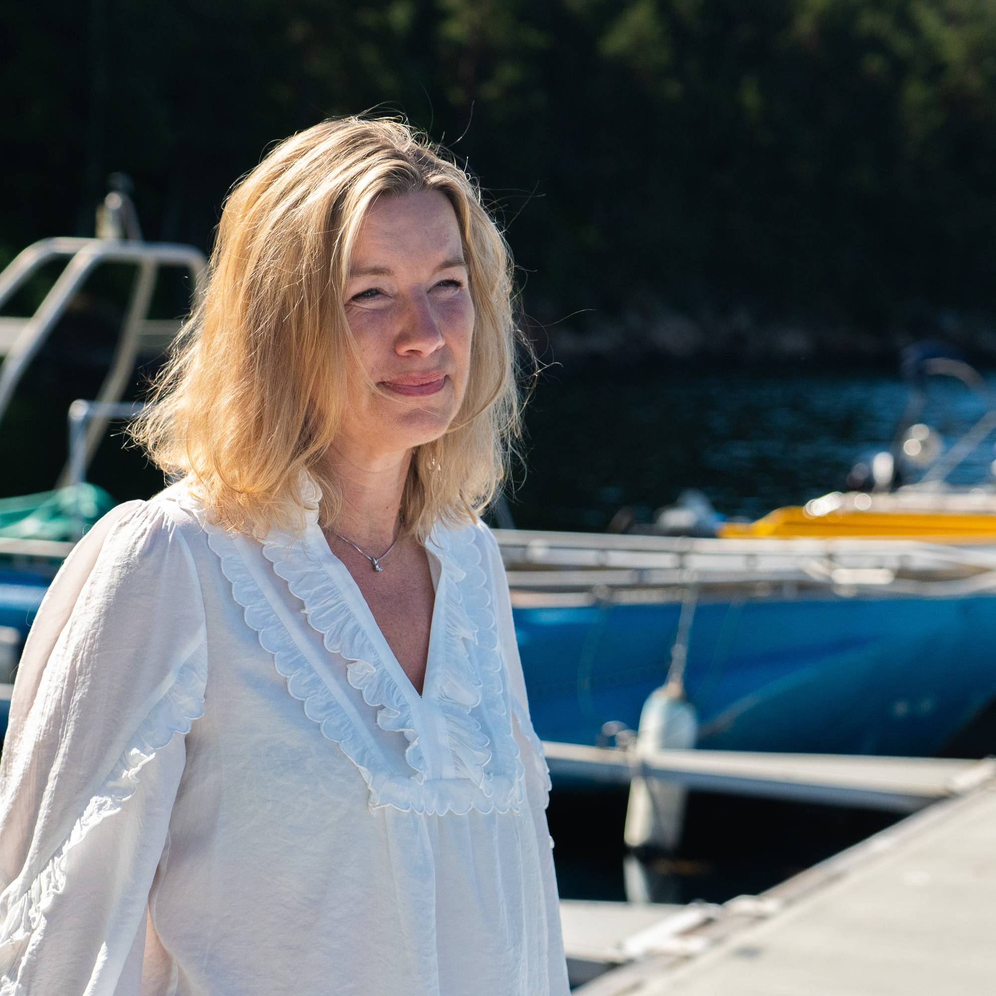 Kvinne med blondt halvlangt hår og hvit bluse står på ei brygge og er utover fjorden