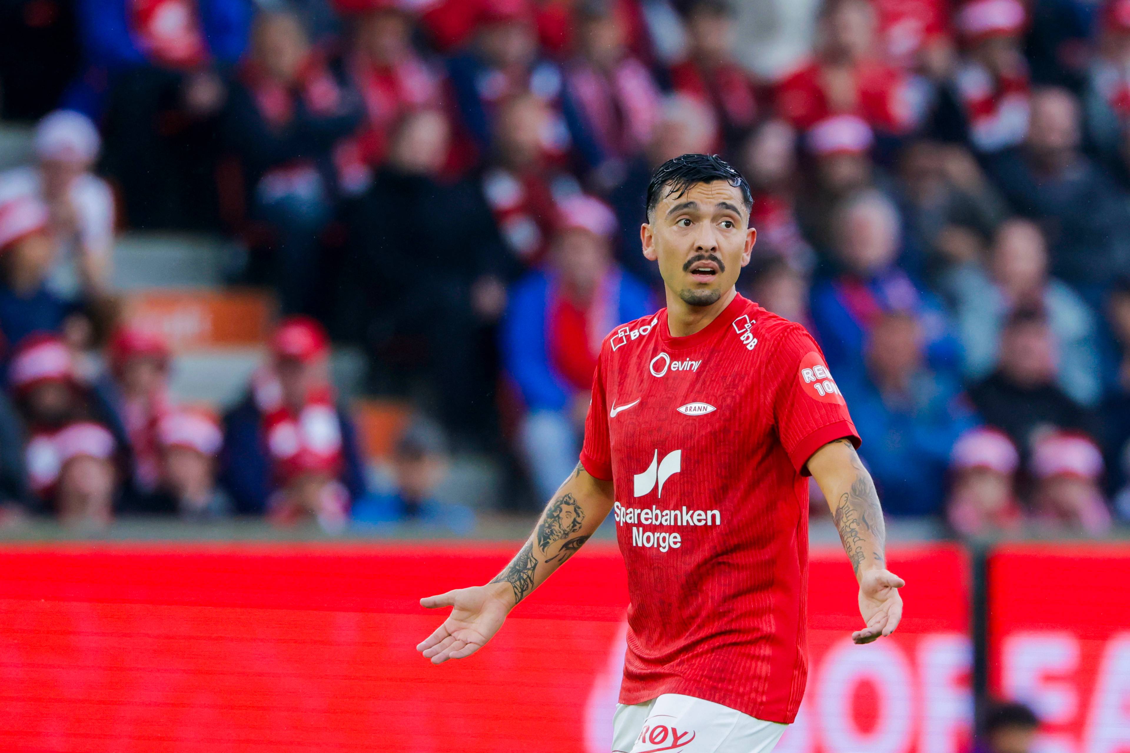 Fotballspiller fra Brann med tatoveringer i rød drakt på banen foran publikum. (Bildebeskrivelsen er laget av en KI-tjeneste)