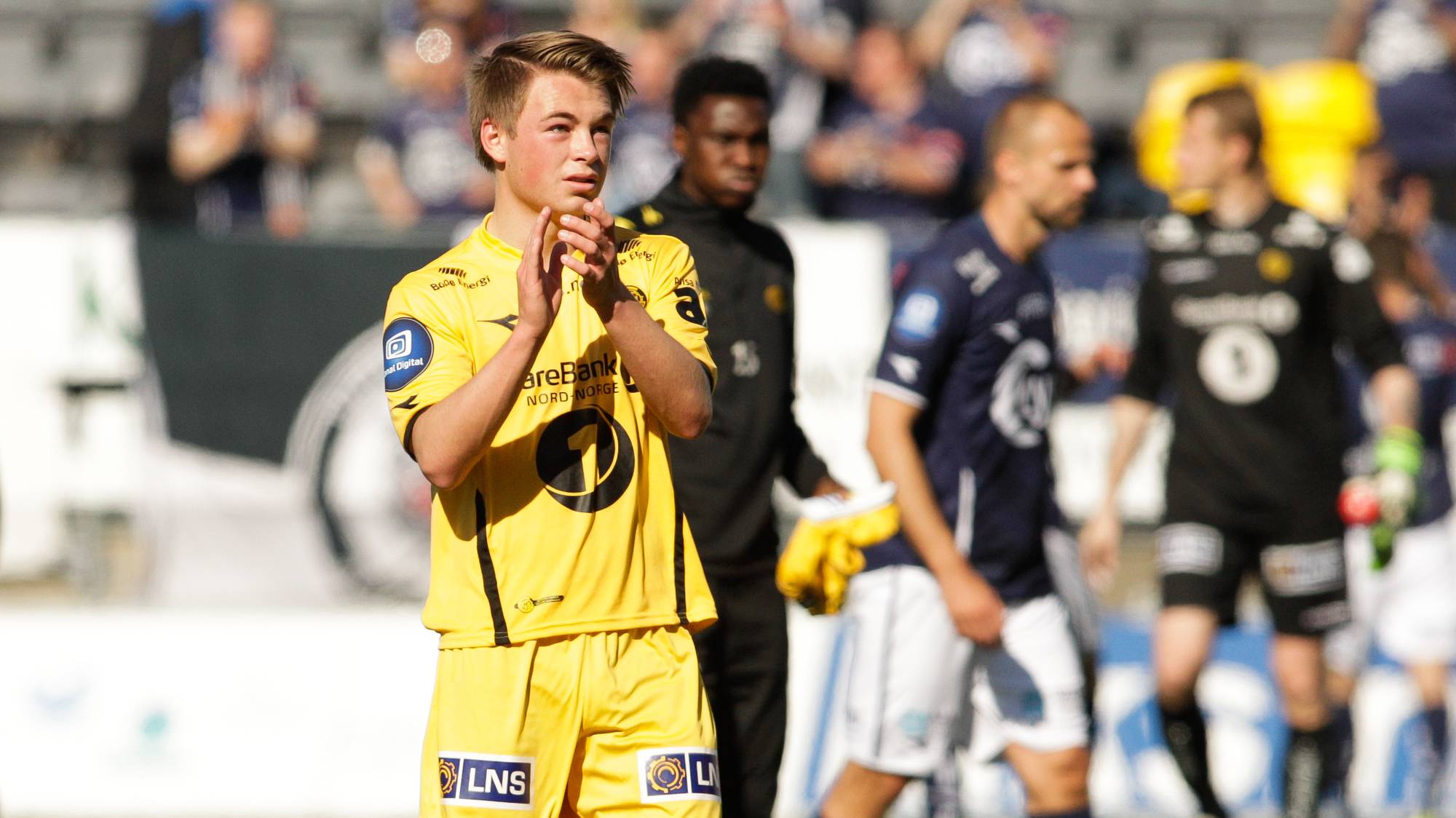 Fotballspiller i gul drakt klapper på banen med andre spillere i bakgrunnen. (Bildebeskrivelsen er laget av en KI-tjeneste)
