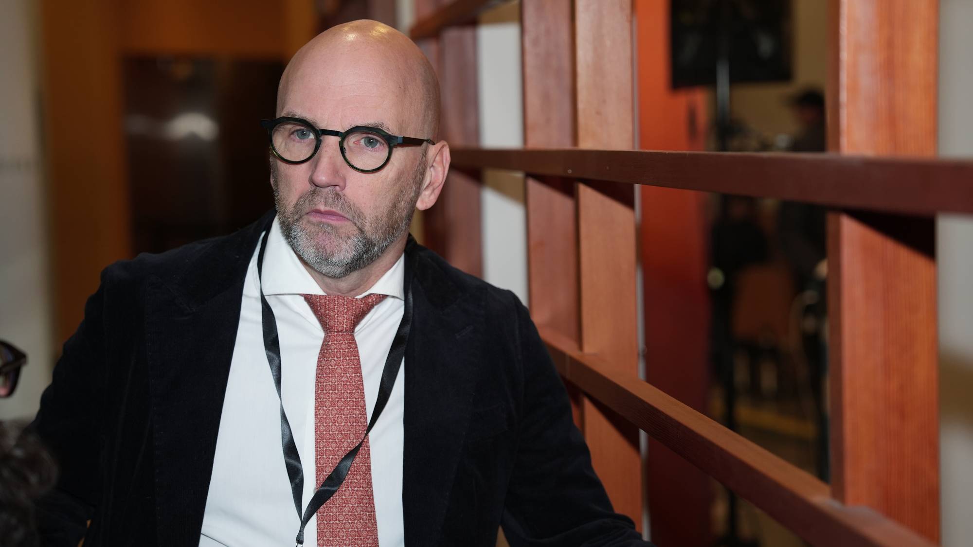 En skallet mann med briller sitter ved siden av et rekkverk av tre. Han har på seg en hvit skjorte, slips og mørk jakke. Mannen har et kort hengende rundt halsen. (Bildebeskrivelsen er laget av en KI-tjeneste)