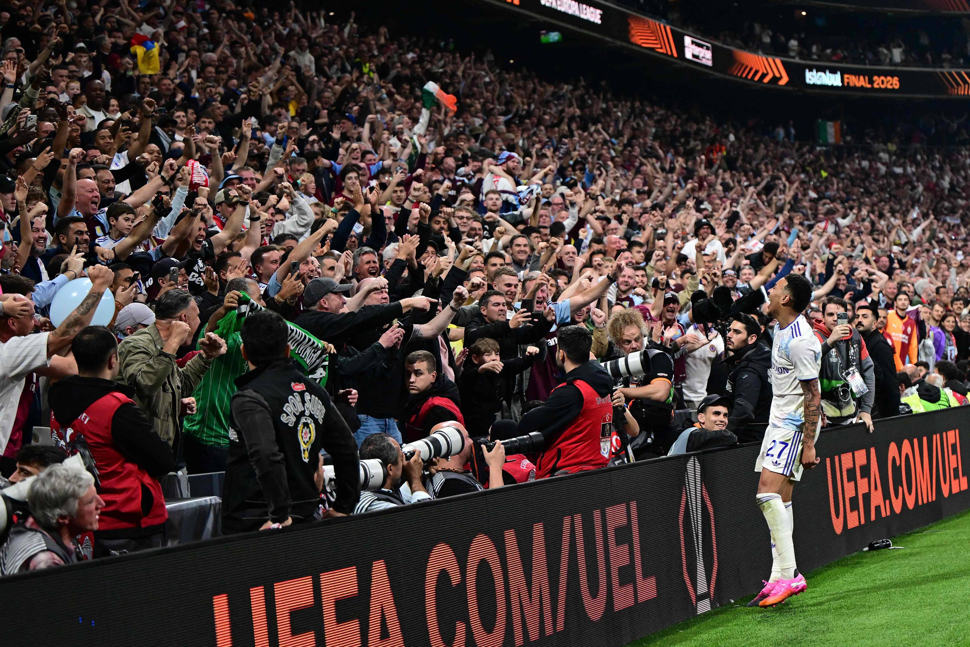 En fotballspiller jubler foran et publikum. Mange tilskuere på tribunen. En stor mengde mennesker hever armene i været. Et UEFA-banner sees i forgrunnen. (Bildebeskrivelsen er laget av en KI-tjeneste)