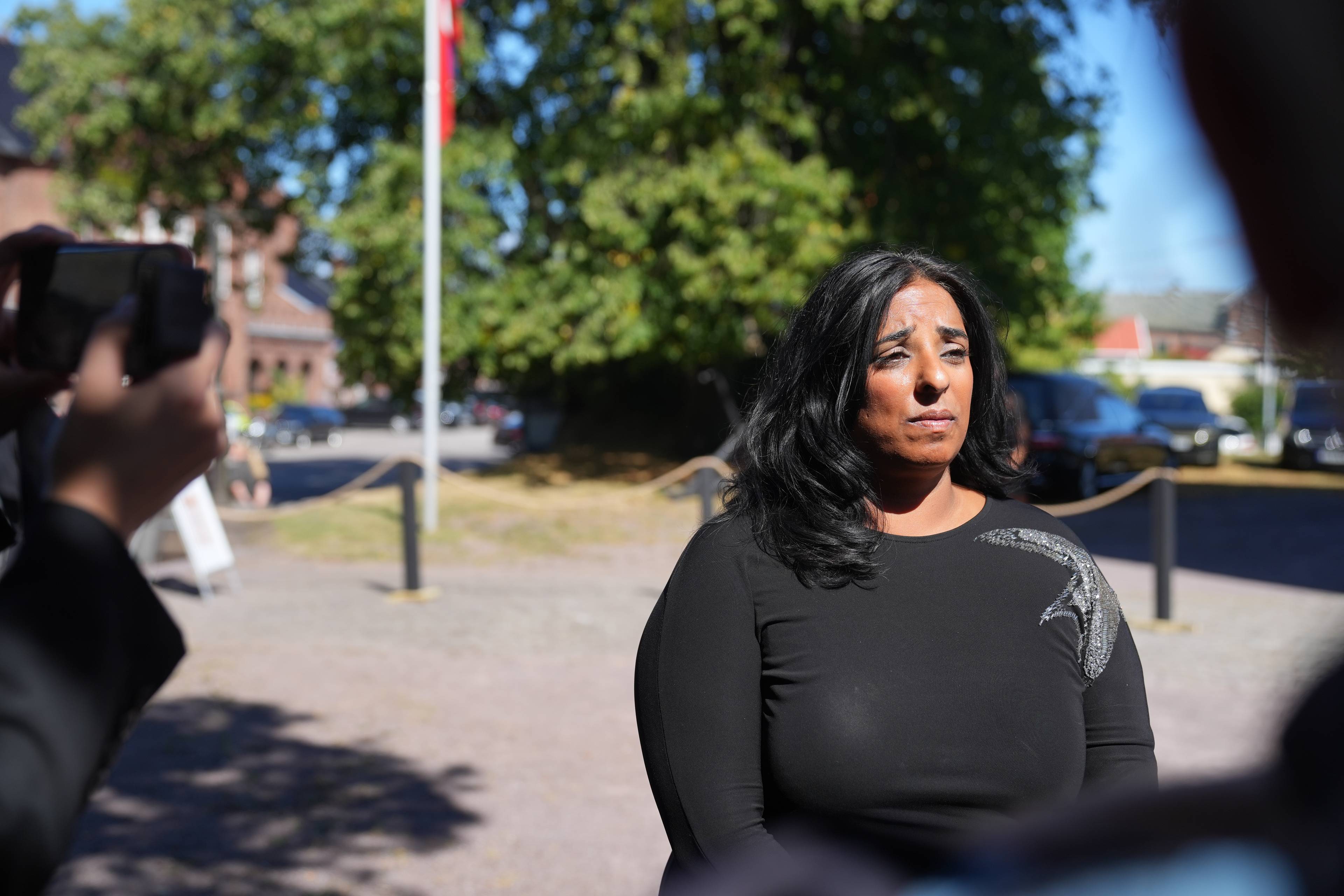 Kvinne i sort kjole intervjues utendørs foran en gruppe pressefolk.