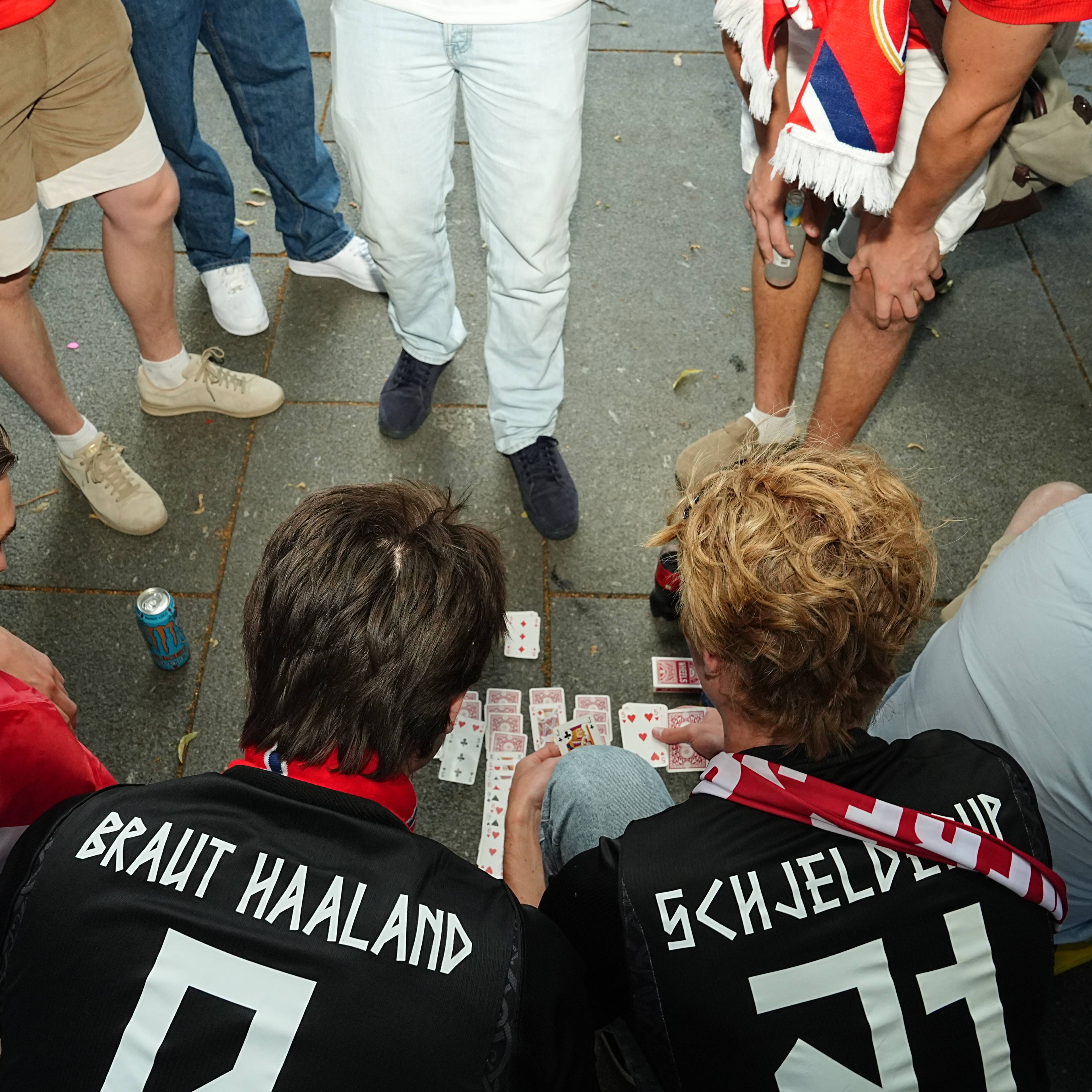 Personer i fotballdrakter sitter på et fortau og spiller kort. (Bildebeskrivelsen er laget av en KI-tjeneste)
