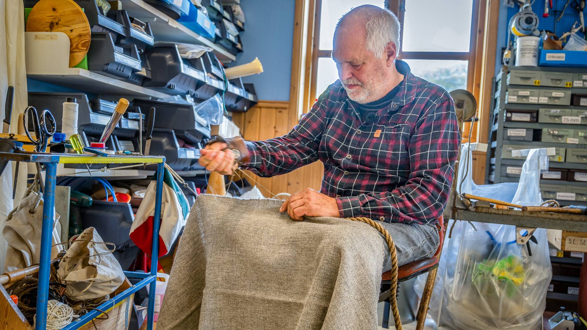 Frode Bjøru, daglig leder på Seglloftet på Jøa. Han sitter på en stol og syr på et seil