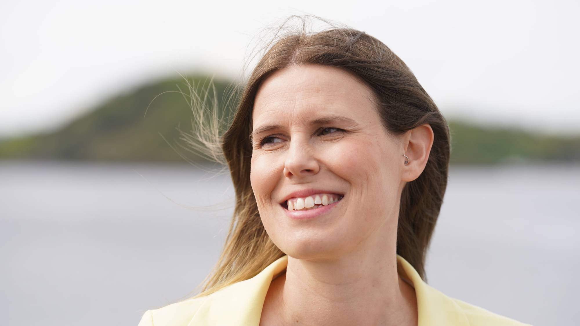 Portrett av en smilende kvinne i gul dress. Hun har brunt hår og ser mot siden. Bak henne er en uskarpt bakgrunn med vann og en åskam. Bildet er tatt utendørs. (Bildebeskrivelsen er laget av en KI-tjeneste)