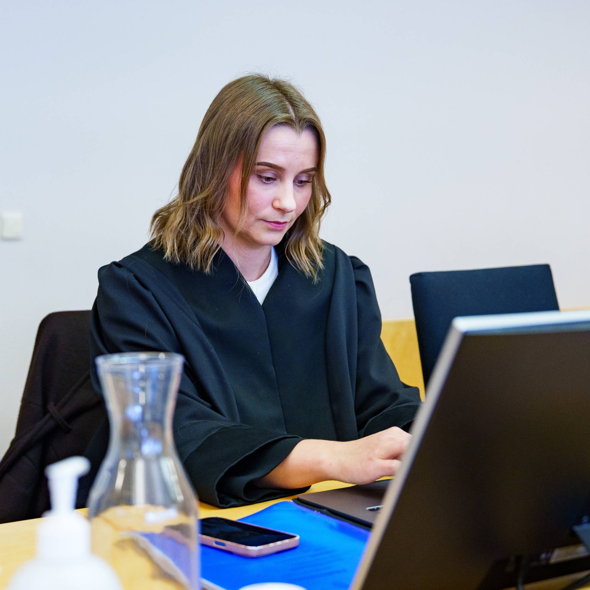 En kvinne i svart advokatkappe sitter ved et skrivebord og jobber på en datamaskin i en rettssal. (Bildebeskrivelsen er laget av en KI-tjeneste)