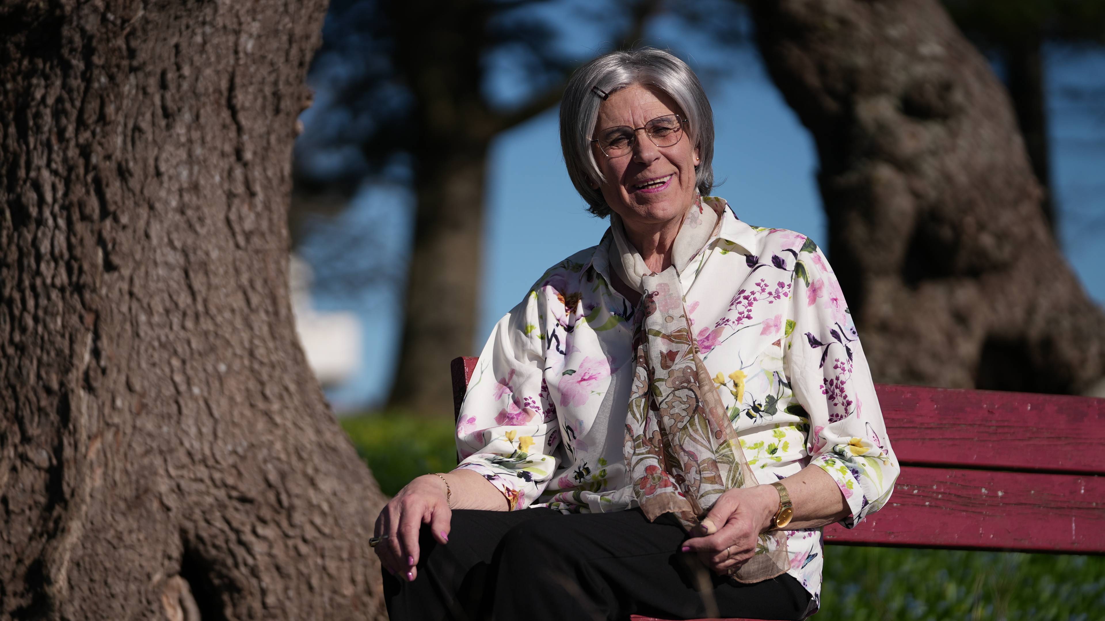 Eldre kvinne sitter på en rød benk ute. Hun har grått hår og briller, og har på seg en blomstrete bluse og sorte bukser. Kvinnen smiler mot kameraet. I bakgrunnen ses trær og en blå himmel. (Bildebeskrivelsen er laget av en KI-tjeneste)