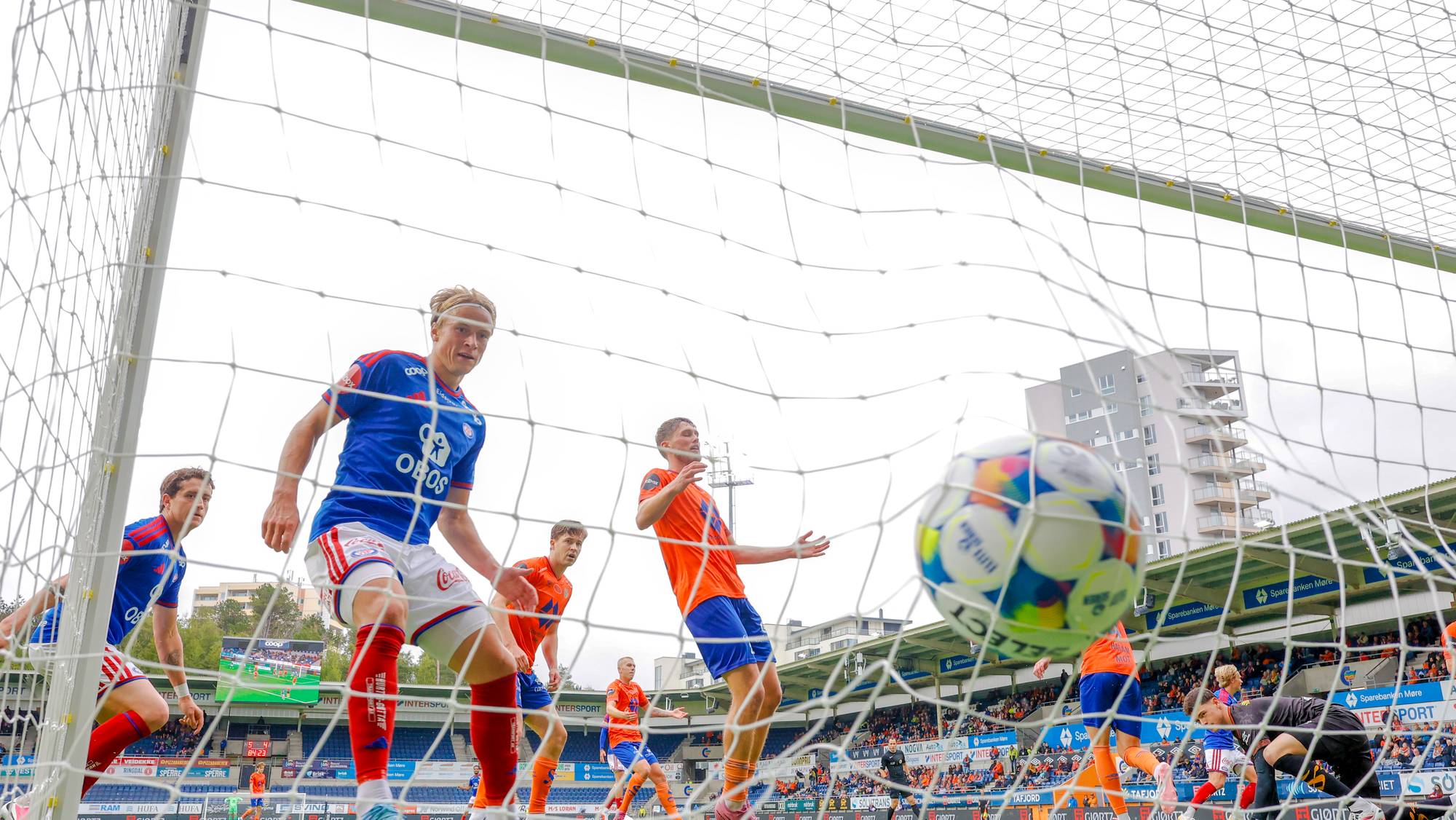 Fotballspillere i en kamp, sett gjennom målet mens ballen er på vei inn. (Bildebeskrivelsen er laget av en KI-tjeneste)