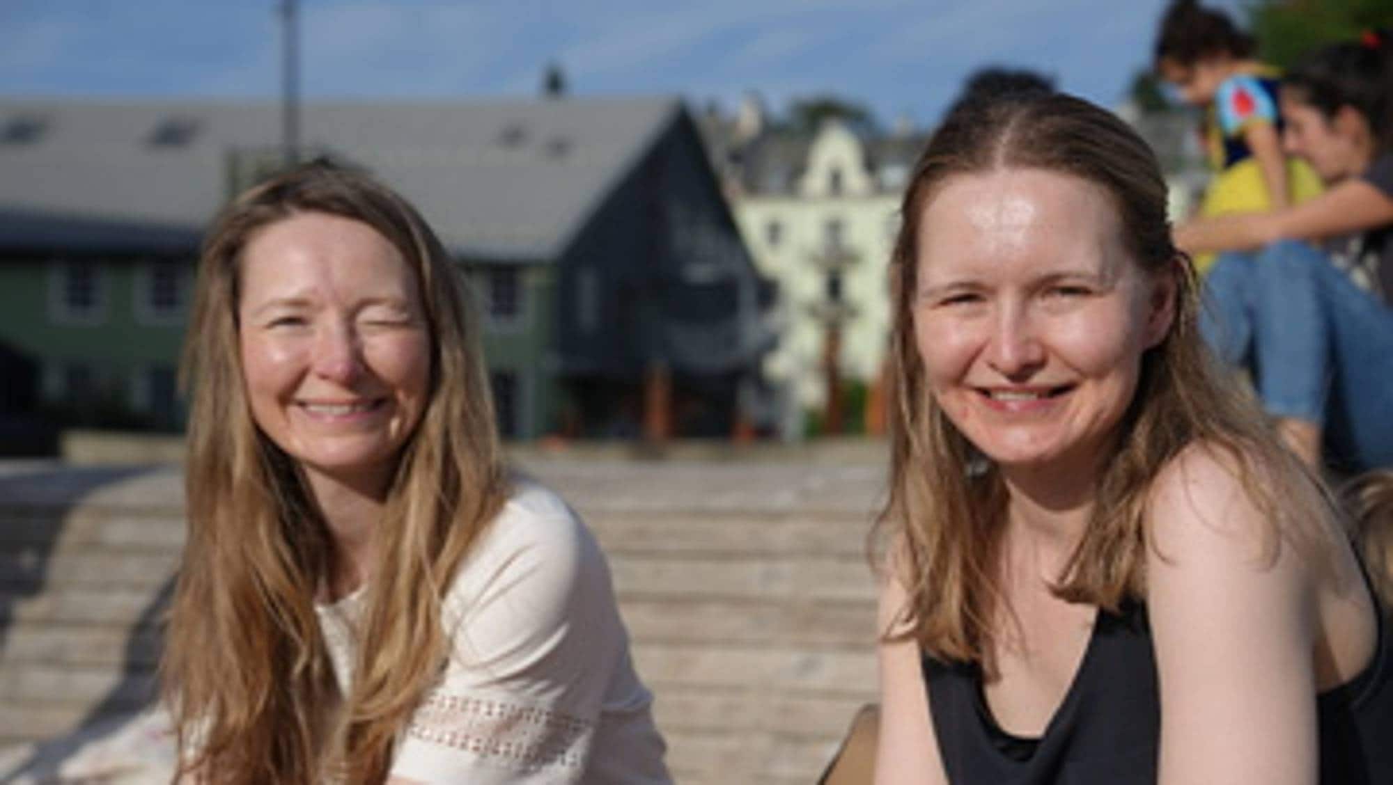 To unge kvinner med høvesvis kvit og svart overdel sit i solskinet og ser i kamera.
