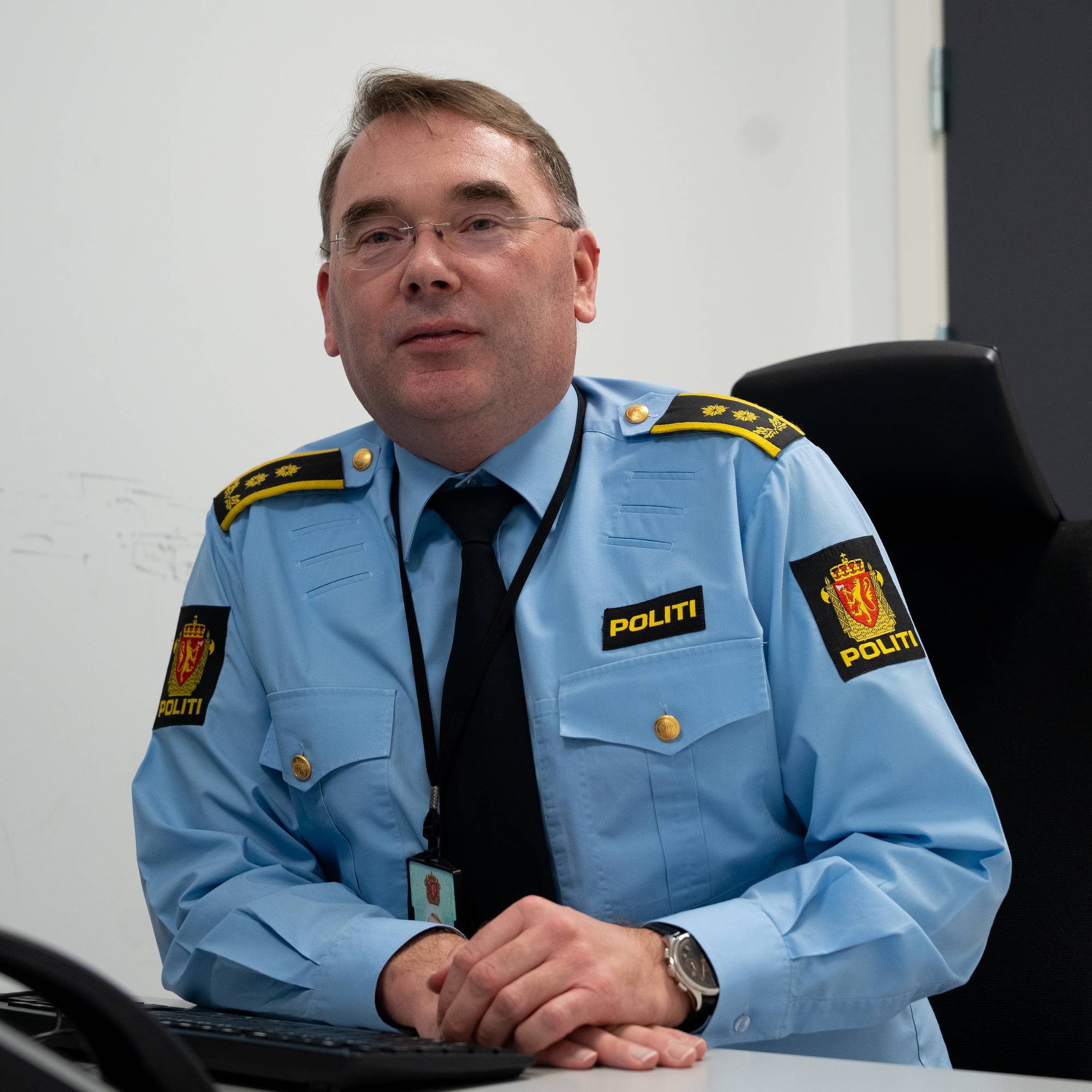 En politibetjent i lyseblå uniform sitter foran en datamaskin. Han har en svart slips og hender plassert på skrivebordet. Mikseren ligger foran ham sammen med en telefon. Bakgrunnen er en nøytral, lysvegg og en dørramme. (Bildebeskrivelsen er laget av en KI-tjeneste)