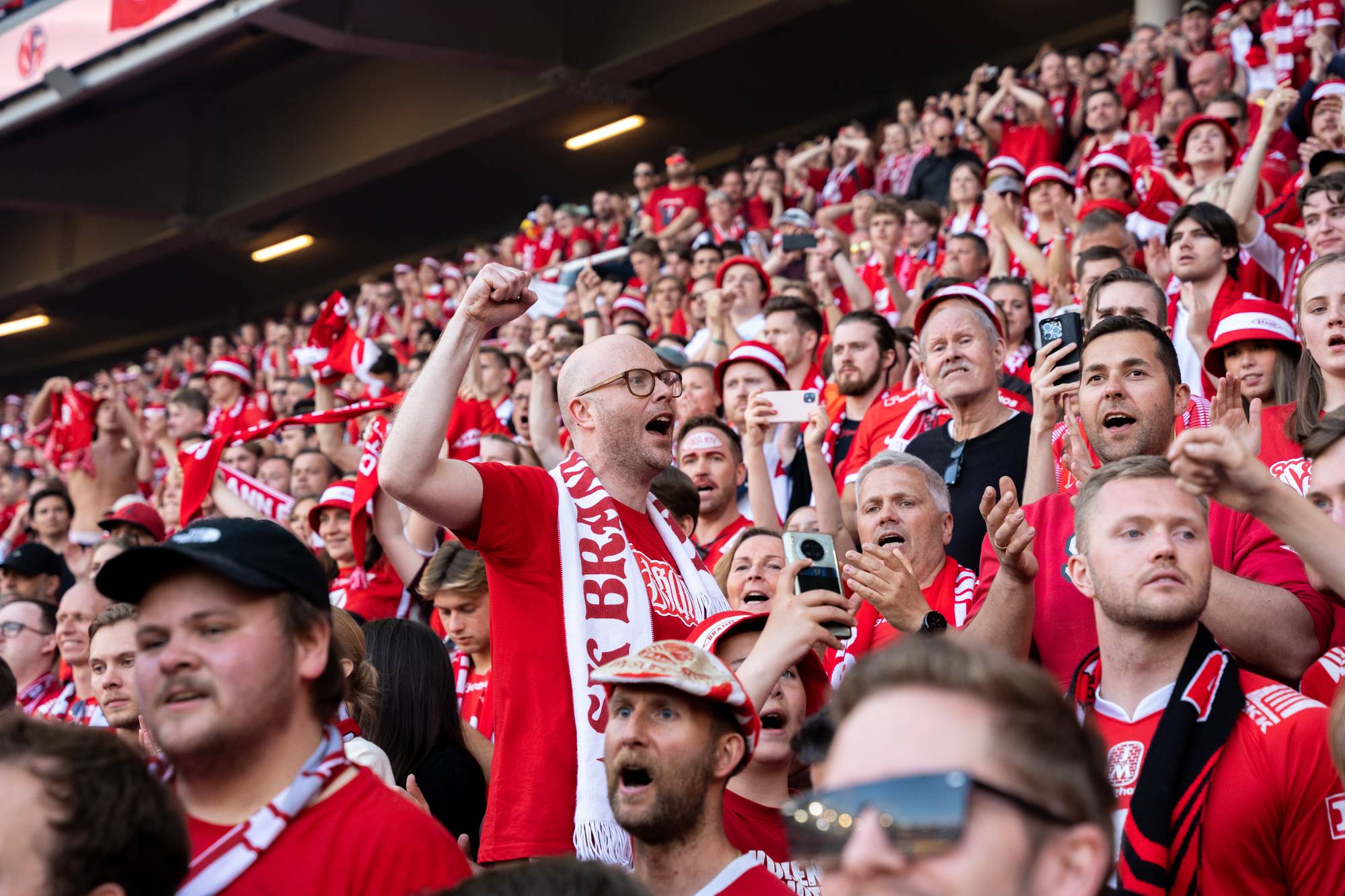 En mann uten hår med briller med Brann-skjerf står i forgrunnen. I bakgrunnen står hundrevis av rød-kledde Brannsupportere på tribunen.