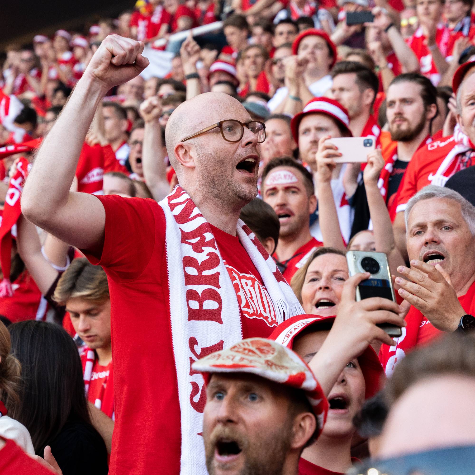 En mann uten hår med briller med Brann-skjerf står i forgrunnen. I bakgrunnen står hundrevis av rød-kledde Brannsupportere på tribunen.