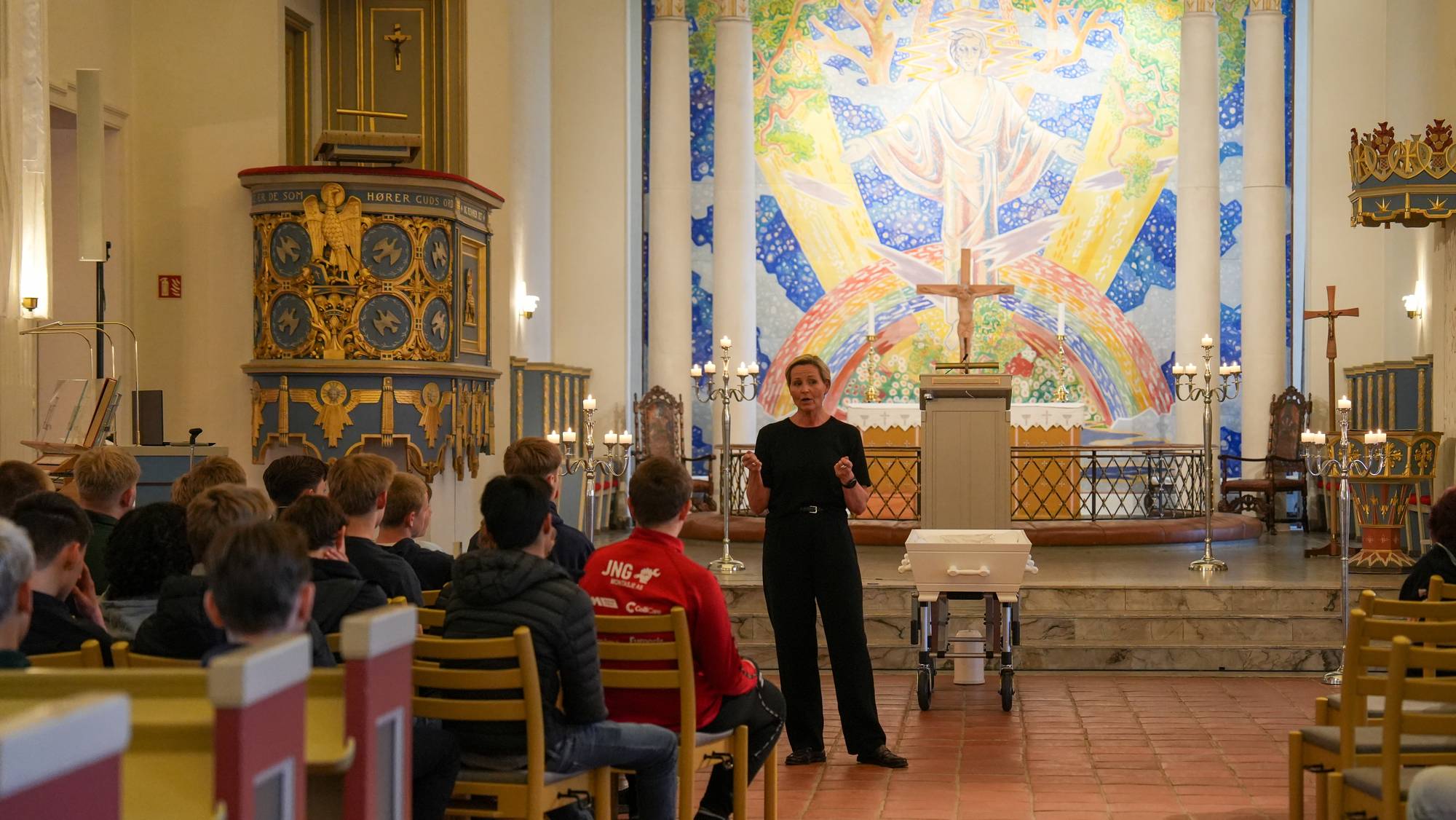 Konfirmasjonskurs i Glemmen kirke. En dame i svart antrekk står foran en gjeng ungdommer og holder kurs. Ved siden av henne er en hvit kiste. I bakgrunnen er det et kirkemaleri. 