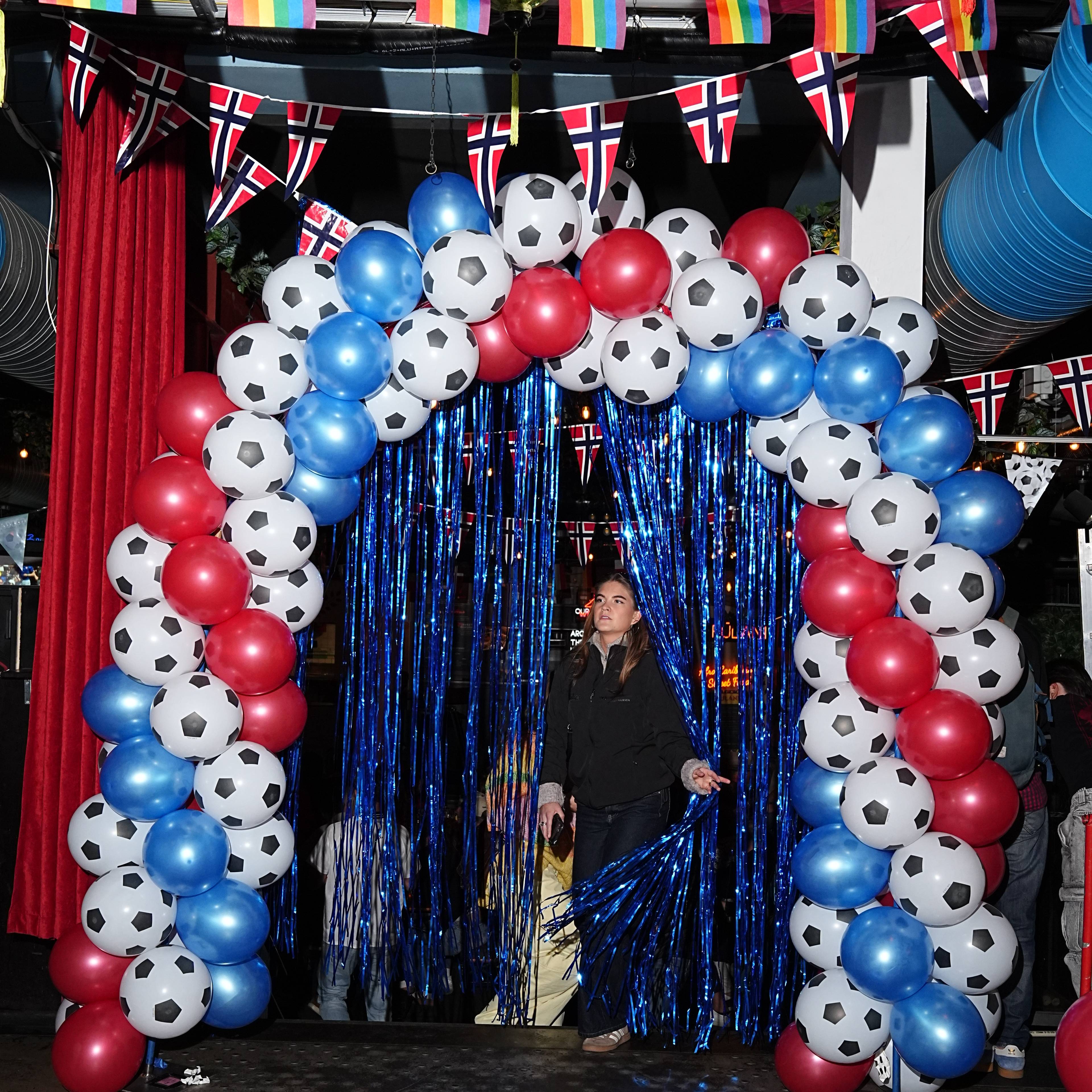En kvinne står bak et blått glitrende forheng, foran en bue laget av fotballformede ballonger. Ballongbuen er dekorert med norske flagg og regnbueflagg. Det er flere personer i bakgrunnen som går bak forhenget. (Bildebeskrivelsen er laget av en KI-tjeneste)