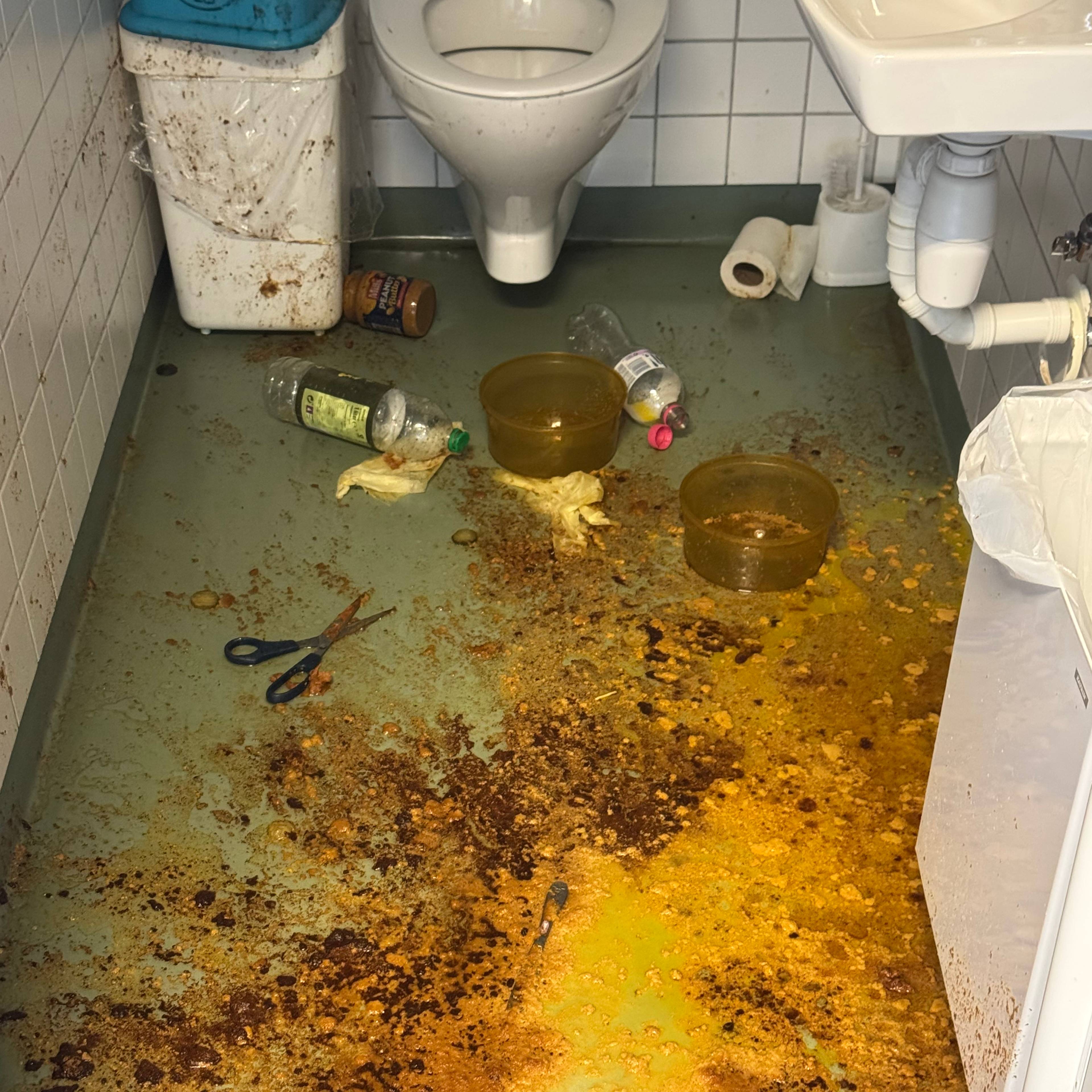 Toalettrom med gulv dekket av søl, avfall, en saks og løse beholdere. (Bildebeskrivelsen er laget av en KI-tjeneste)