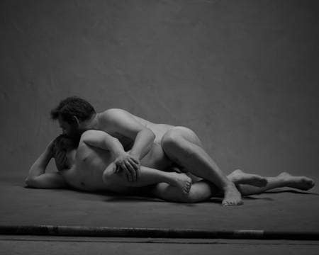 To nakne menn ligger bak hverandre ei skje på gulvet mens de kysser