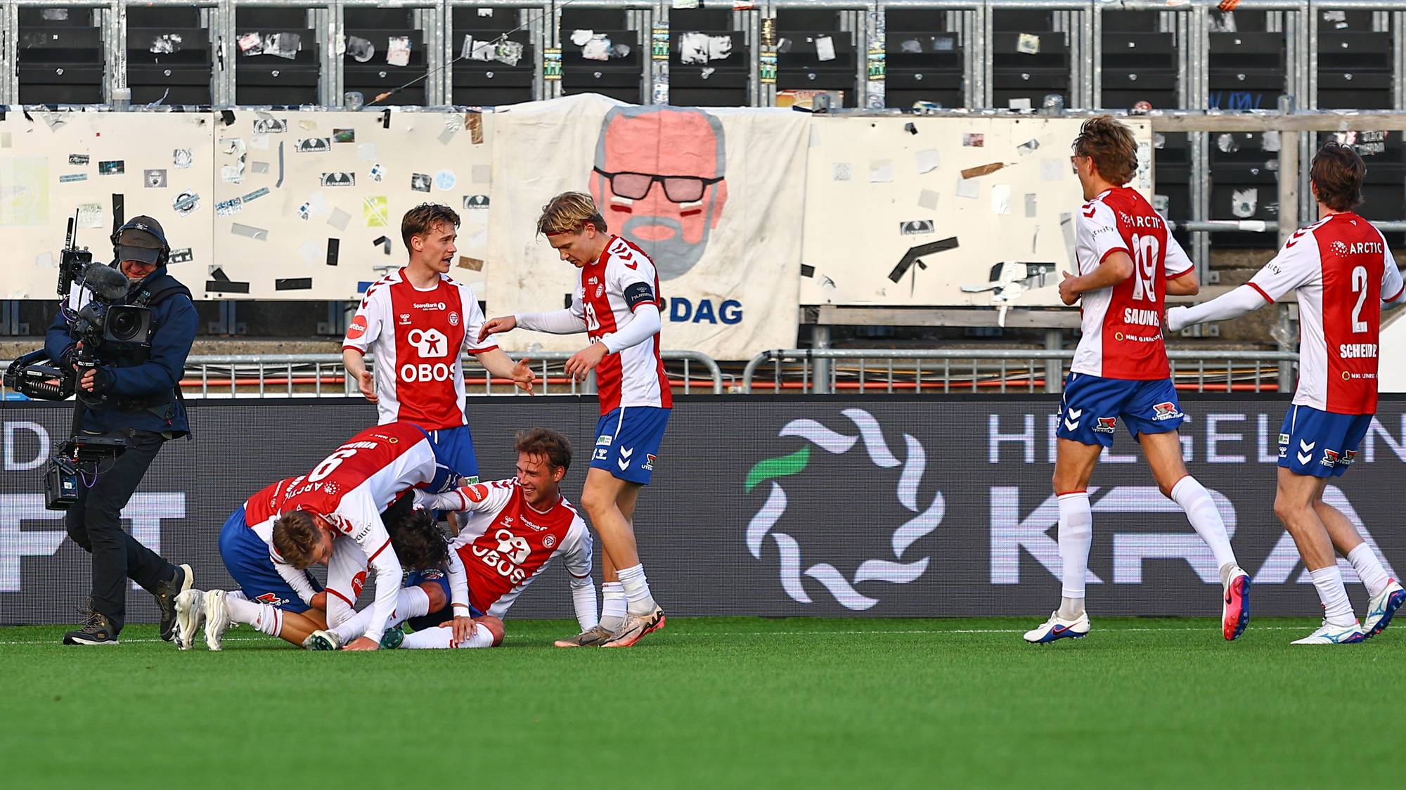Fotballspillere feirer på en gressbane. Flere spillere ligger nede og blir gratulert av lagkamerater. En kameramann filmer hendelsen. I bakgrunnen er tribuner med seterekker. (Bildebeskrivelsen er laget av en KI-tjeneste)