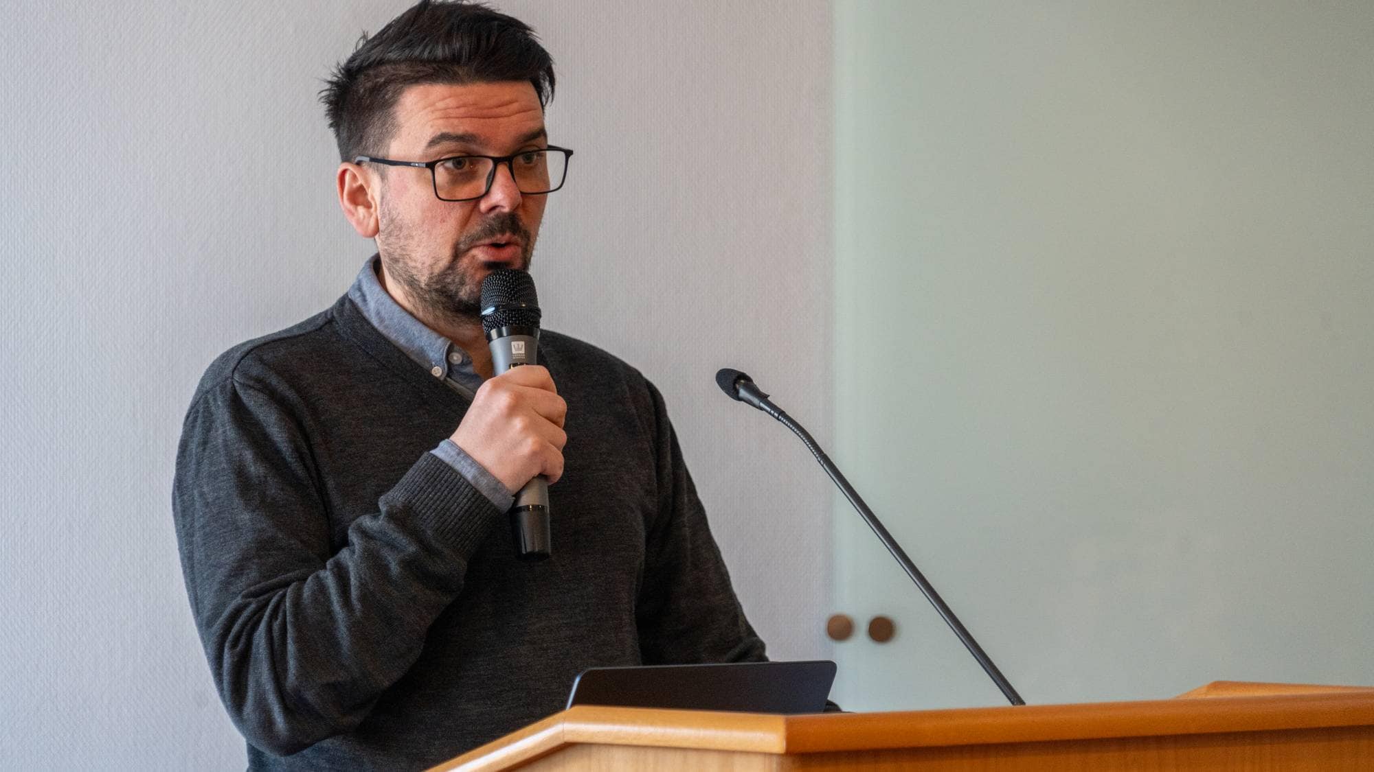 En mann står bak et podium og holder en mikrofon. Han har briller og er iført en mørk genser. Omgivelsene er en lys konferanserom med en enkel vegg i bakgrunnen. Det er en laptop plassert på podiet foran ham. (Bildebeskrivelsen er laget av en KI-tjeneste)