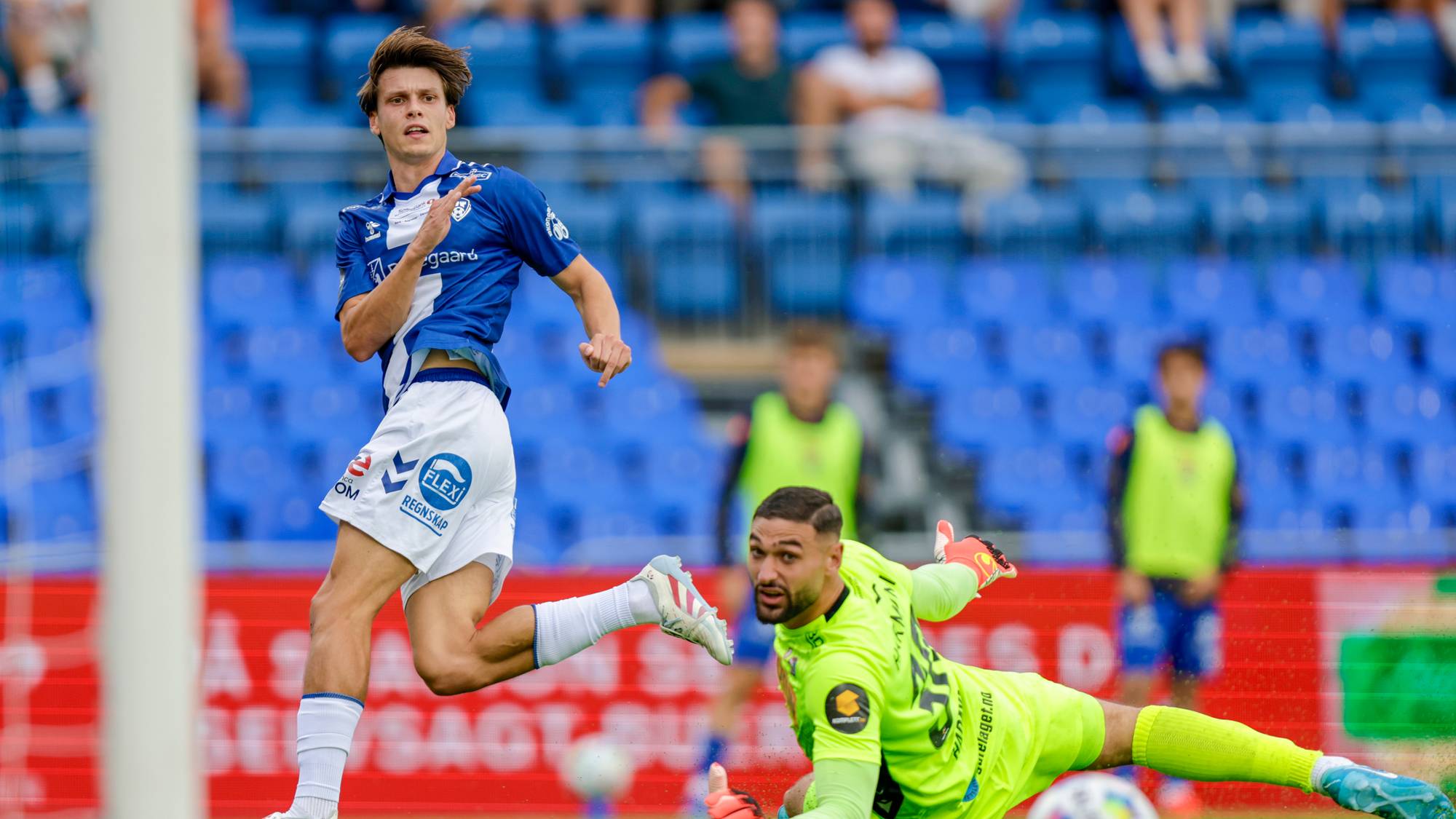 Fotballspiller i blå drakt runder keeper i gul drakt foran mål på en fotballstadion. (Bildebeskrivelsen er laget av en KI-tjeneste)