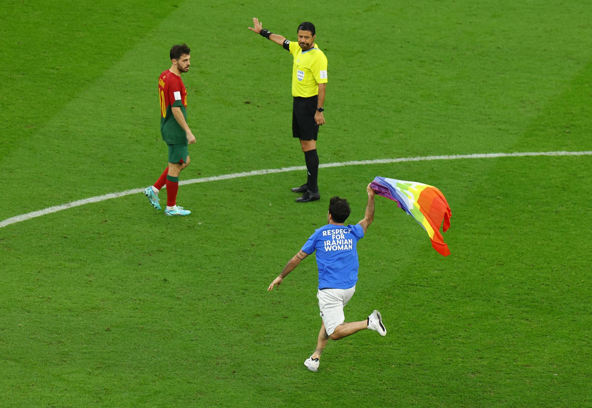 En person med regnbueflagg løper inn på fotballbanen foran en dommer og en spiller. (Bildebeskrivelsen er laget av en KI-tjeneste)