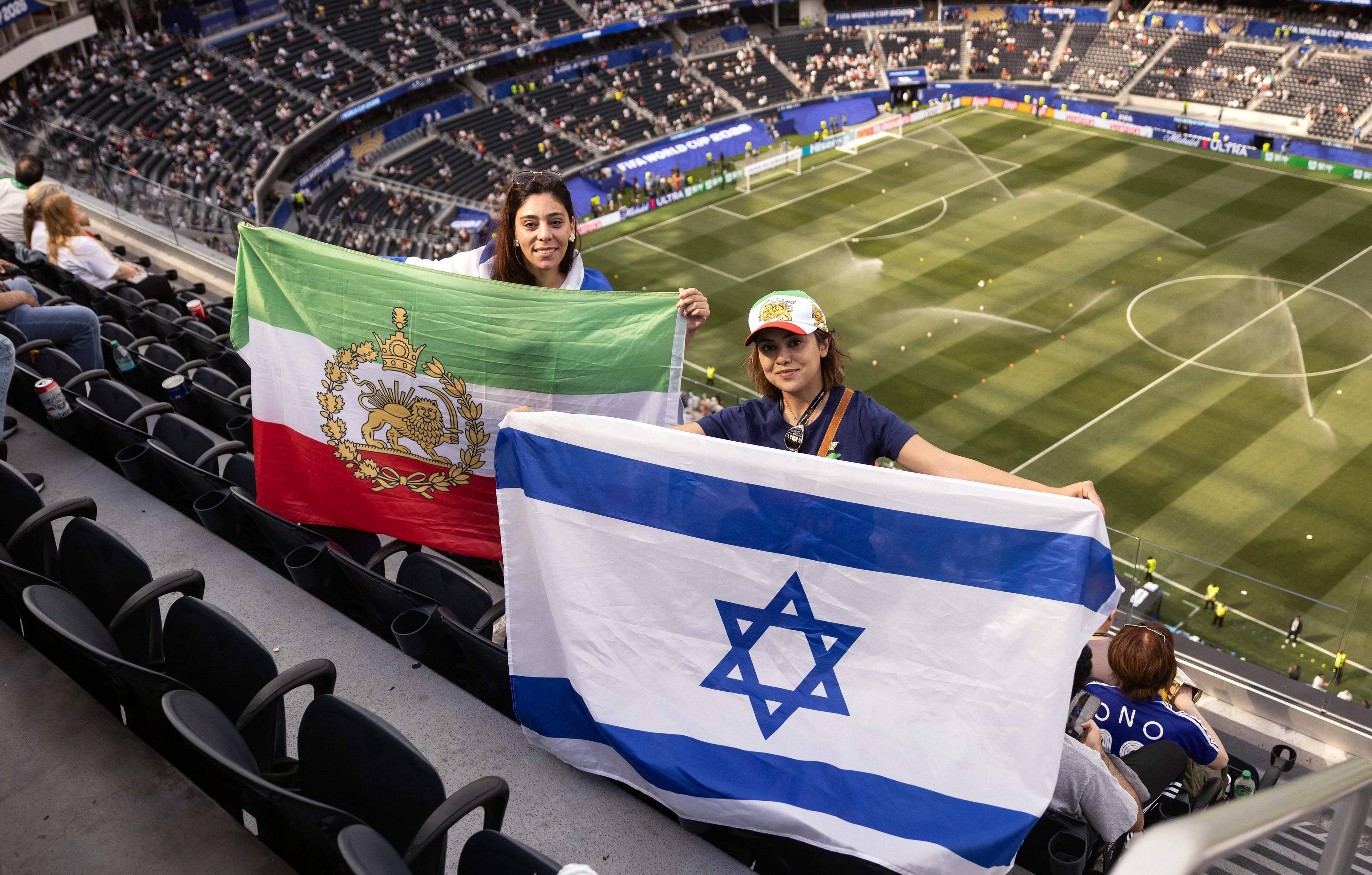 To kvinner holder iranske og israelske flagg på en fotballstadion. De sitter på tribunen med en fotballbane og publikum i bakgrunnen. Begge kvinnene smiler mot kamera. (Bildebeskrivelsen er laget av en KI-tjeneste)