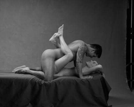 En naken mann med tatoveringer og mørkt hår ligger over en naken kvinne som har bena rundt ryggen hans