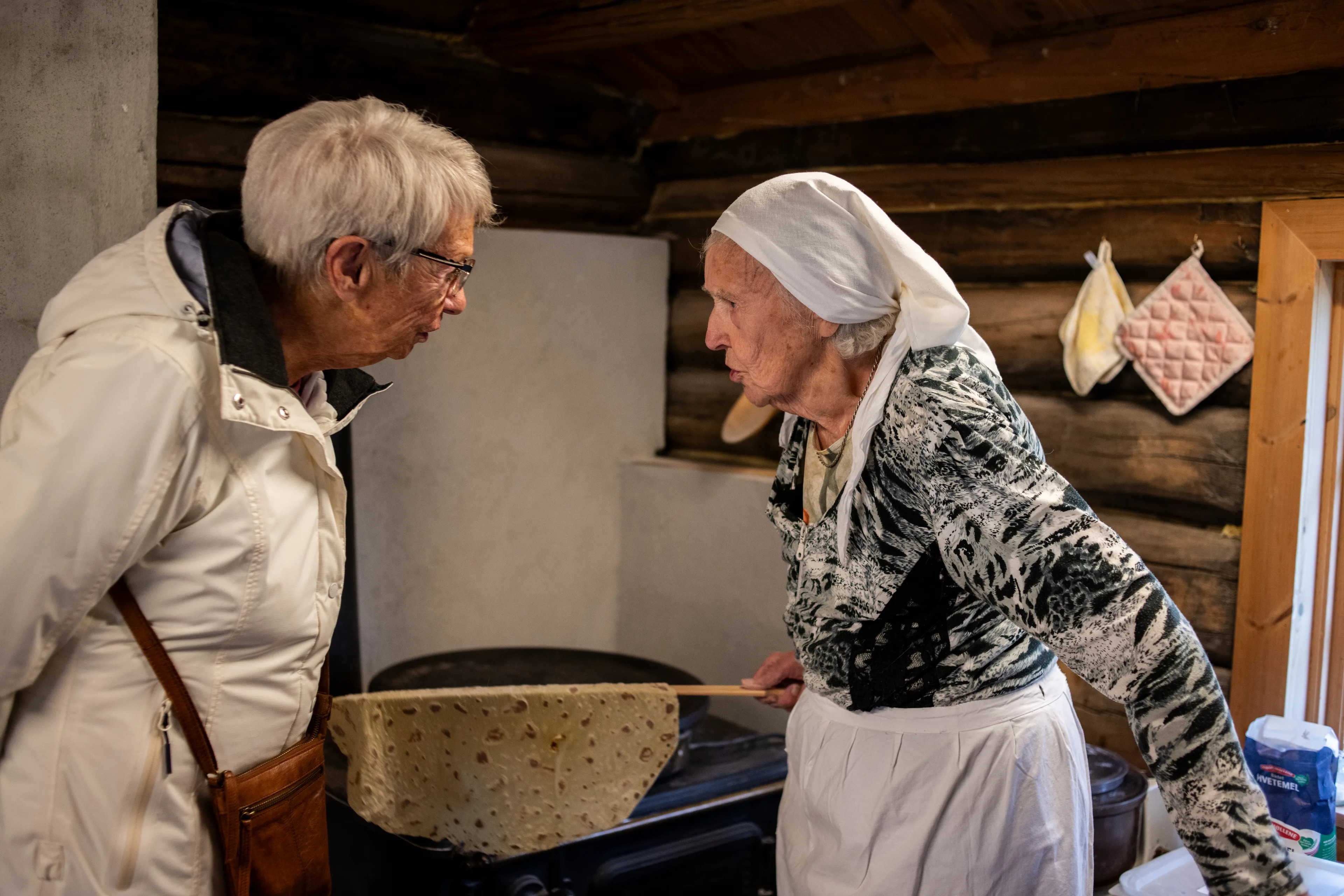 To eldre damer ser på hverandre, den ene holder en takkestav med potetlefse