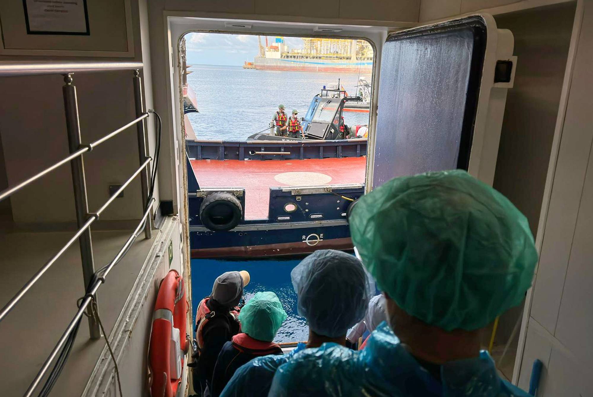 Personer kledd i smitteverndrakter står på en døråpning og ser ut på sjøen. Utenfor er et skip, en liten båt og en oljeplattform i bakgrunnen.