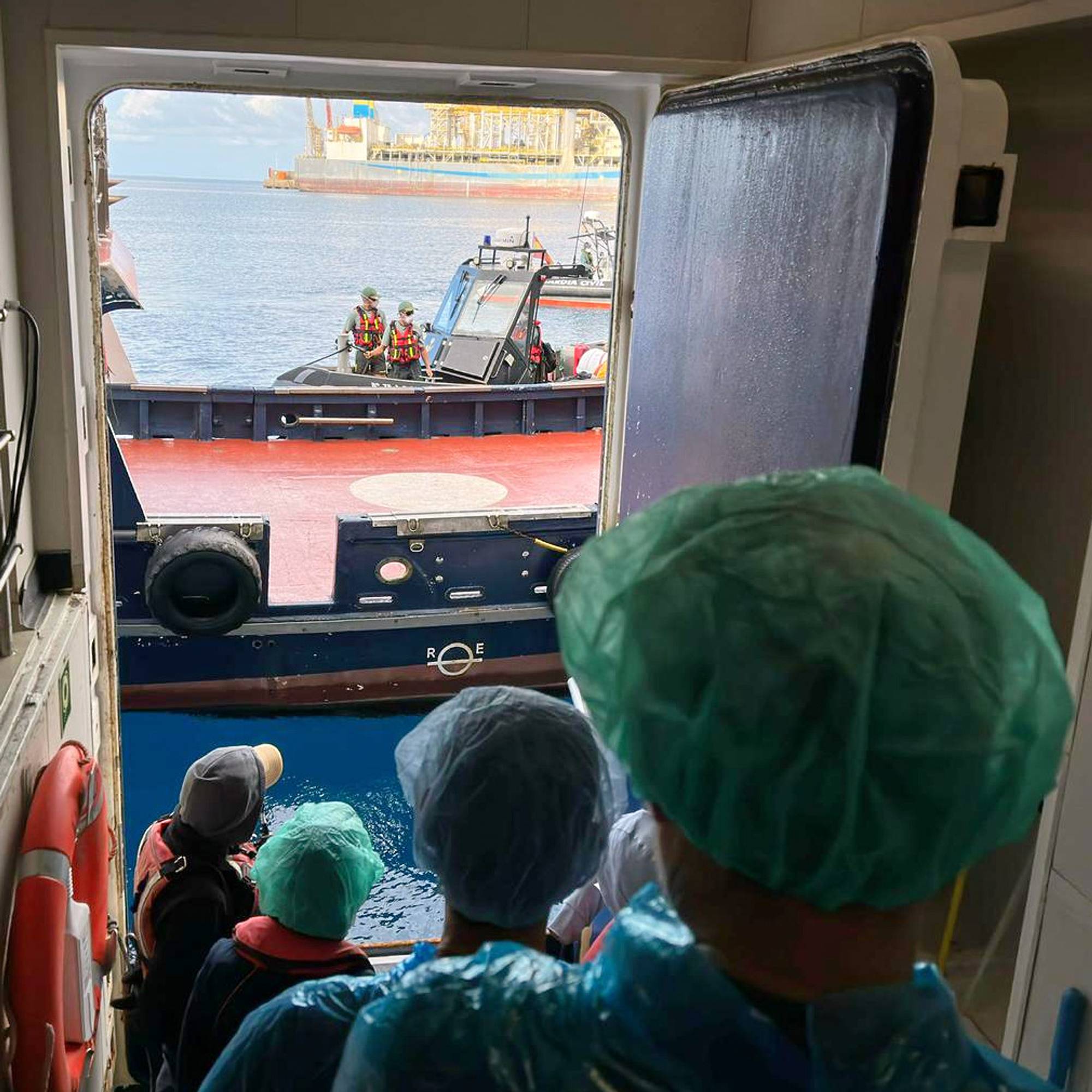Personer kledd i smitteverndrakter står på en døråpning og ser ut på sjøen. Utenfor er et skip, en liten båt og en oljeplattform i bakgrunnen.