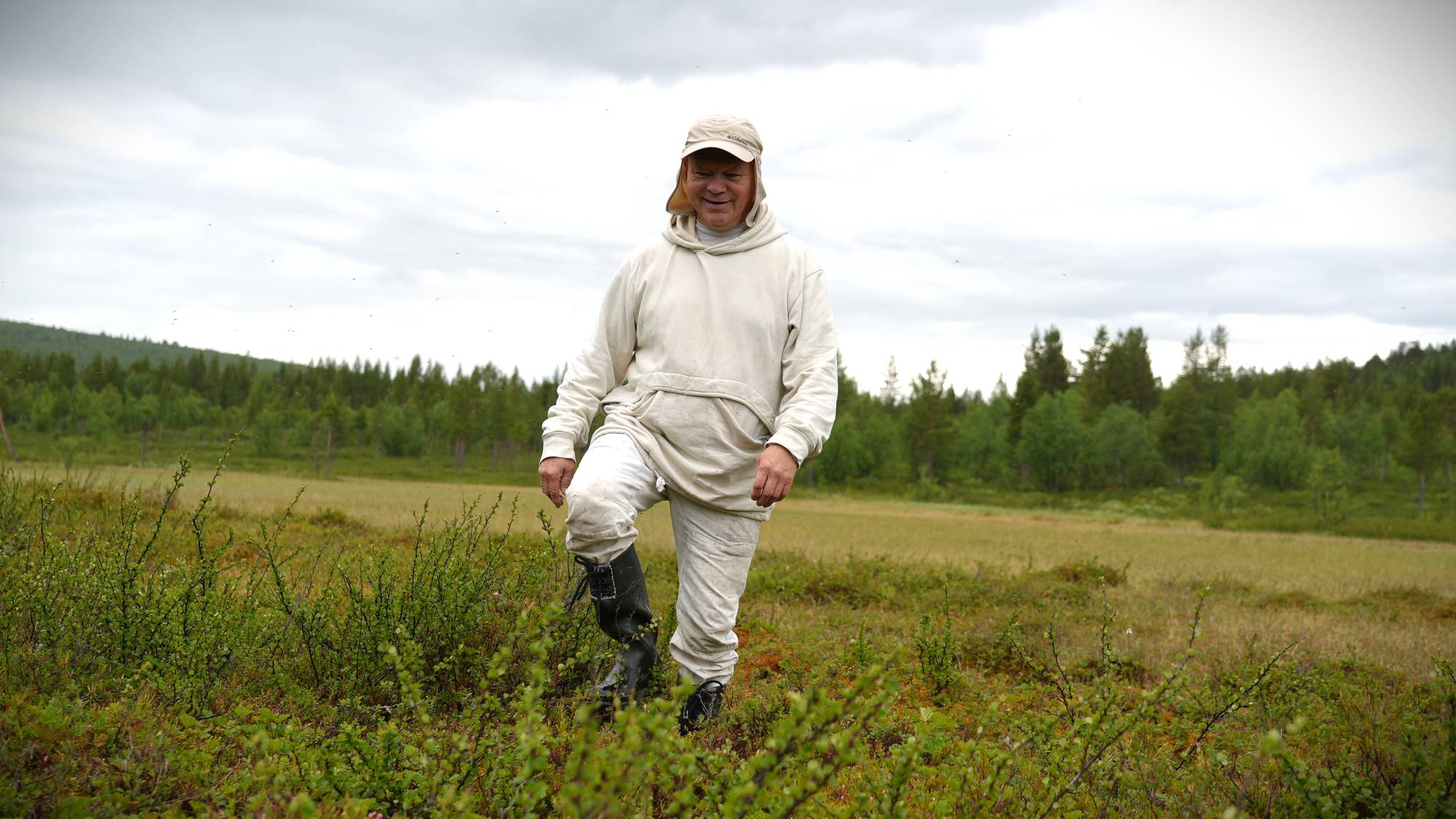 En mann med hvit hettegenser, bukse og kaps, og svarte støvler går i en myr.
