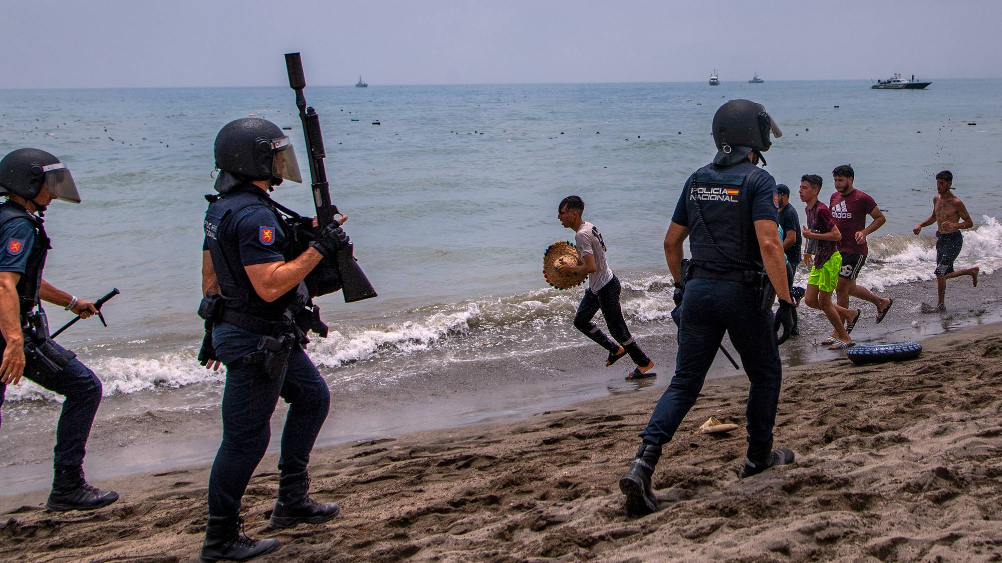 Spansk politi møter unge menn på en strand. Bakgrunnen viser hav og båter i horisonten. (Bildebeskrivelsen er laget av en KI-tjeneste)