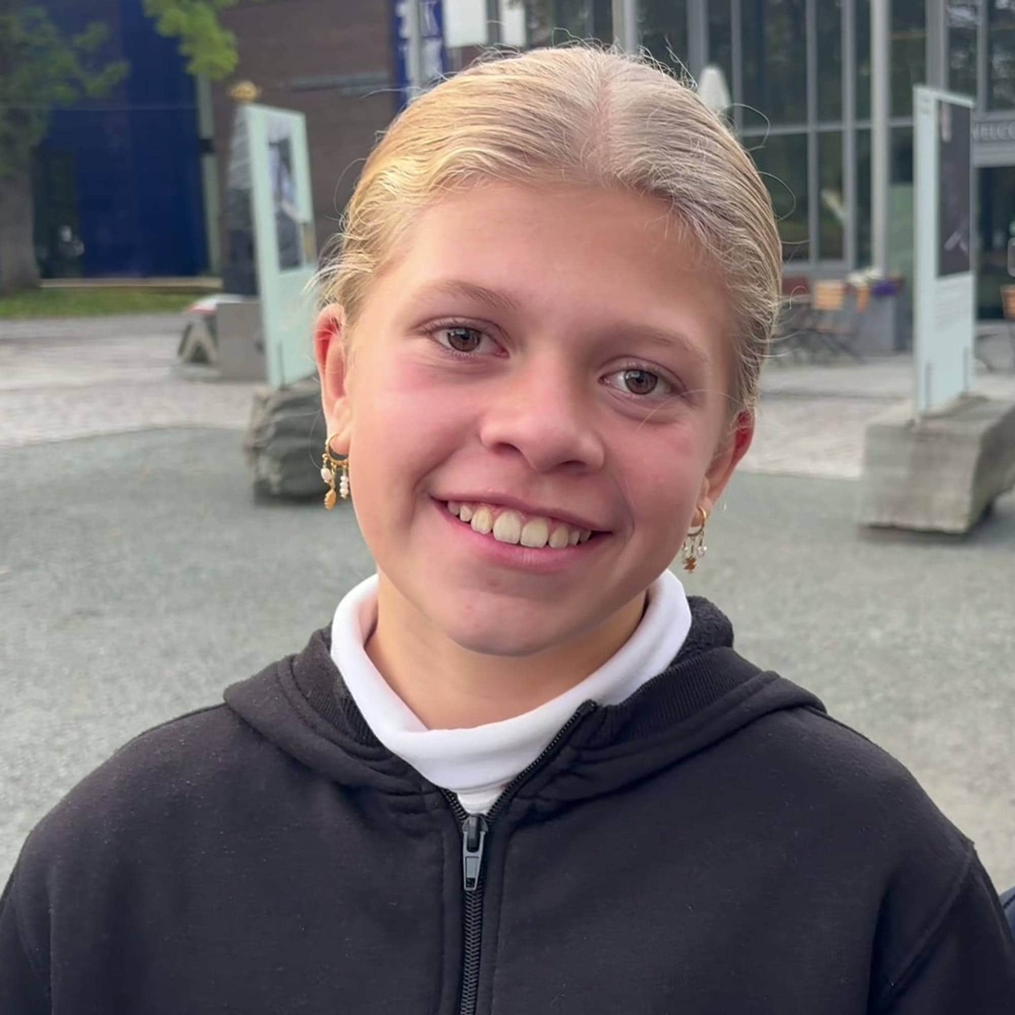 To unge jenter smiler til kamera utendørs ved en bygning. En kvinne står i bakgrunnen. (Bildebeskrivelsen er laget av en KI-tjeneste)