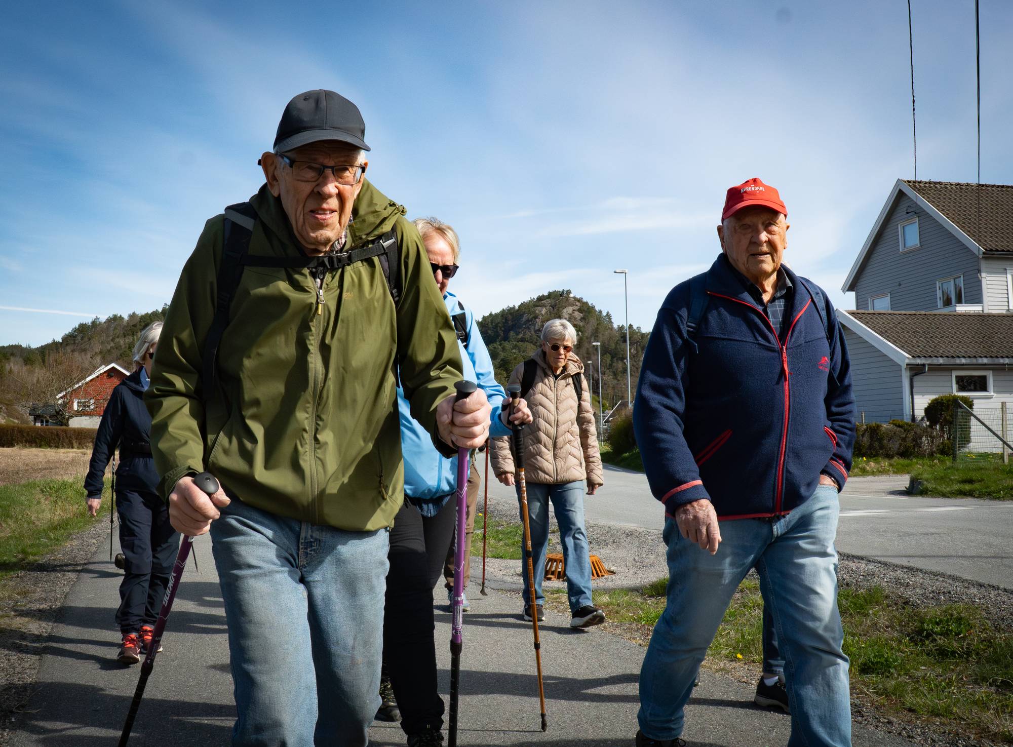 En gruppe eldre mennesker går en tur med turstaver. Mennene og kvinnene er kledd for utendørsaktivitet i uformelle klær. De går langs en vei med hus og vegetasjon i bakgrunnen. (Bildebeskrivelsen er laget av en KI-tjeneste)