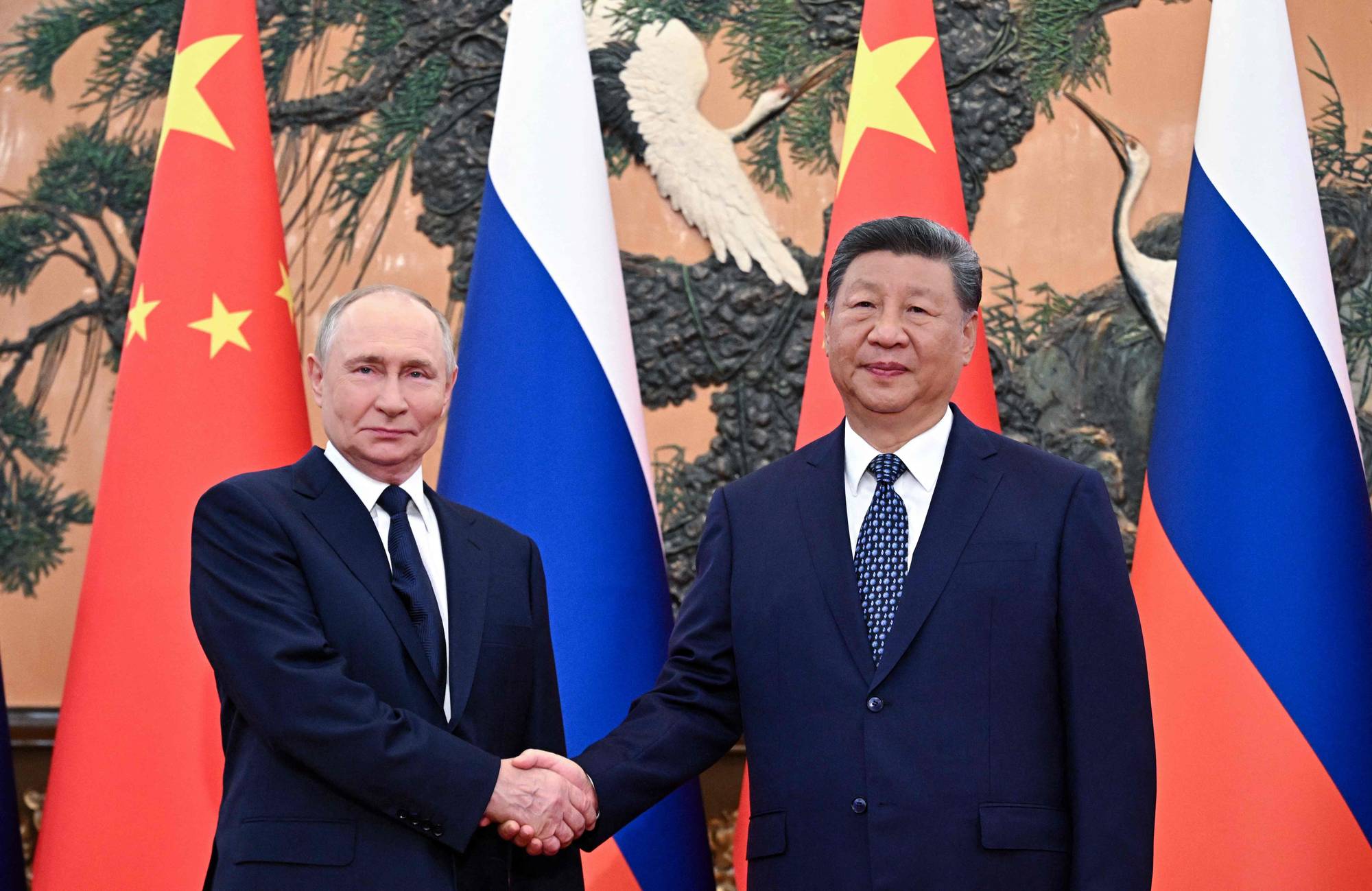 Vladimir Putin og Xi Jinping står ved siden av hverandre foran flaggene til Kina og Russland. De to lederne hilser hverandre med håndtrykk. (Bildebeskrivelsen er laget av en KI-tjeneste)