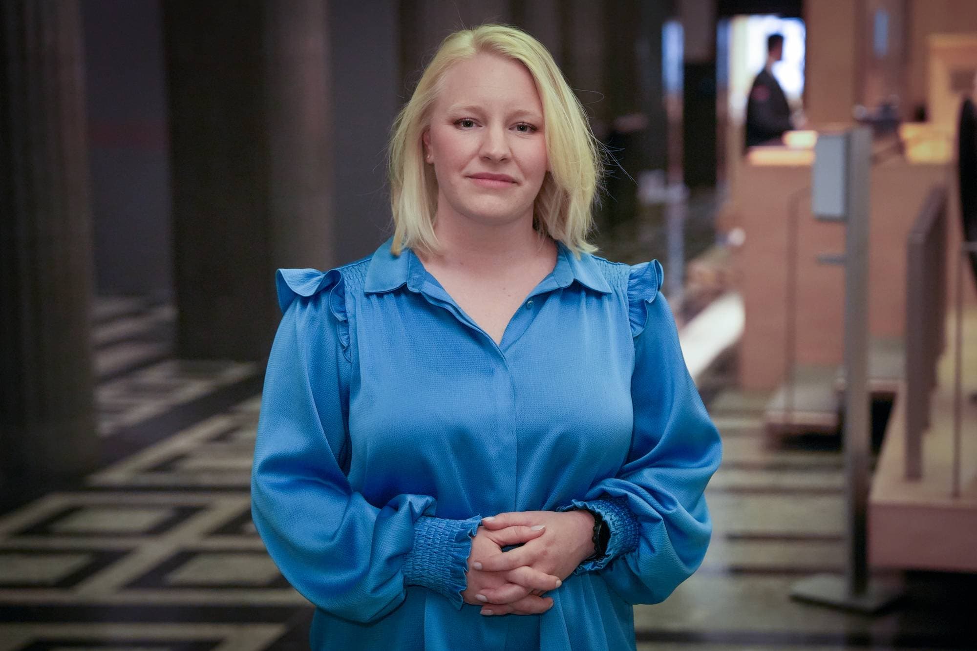 En kvinne med blondt hår i blå kjole står og ser mot kamera. Hender er knyttet foran henne. I bakgrunnen sees en person i uniform. 