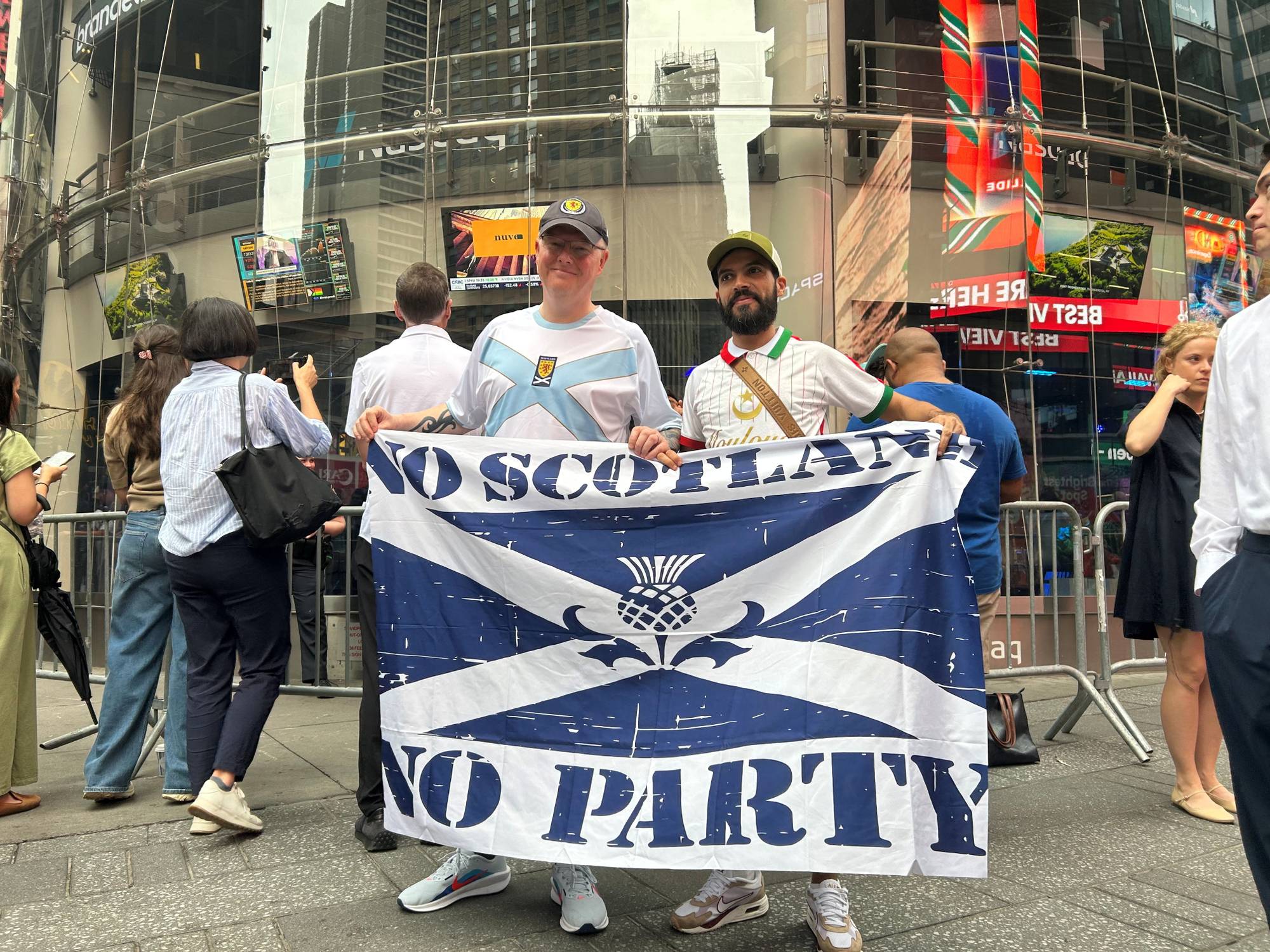 To menn holder et stort skotsk flagg på et travelt bytorv. Flagget har teksten "NO SCOTLAND NO PARTY". Flere personer er synlige i bakgrunnen, og det er store skjermer på bygningene. (Bildebeskrivelsen er laget av en KI-tjeneste)