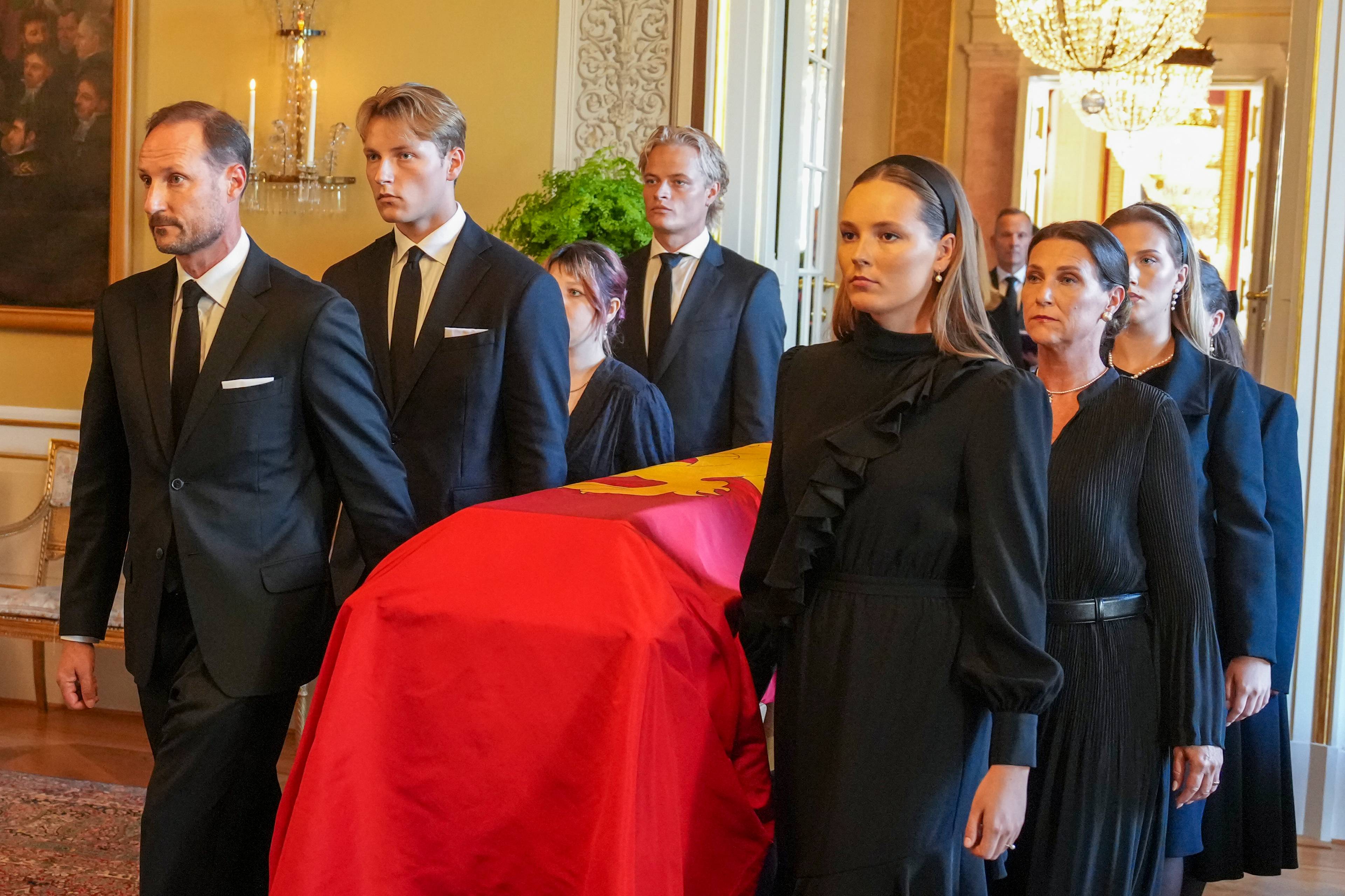 Kong Haakon og kongefamilien går i begravelsesfølge med en kiste drapert i rødt og gull. (Bildebeskrivelsen er laget av en KI-tjeneste)