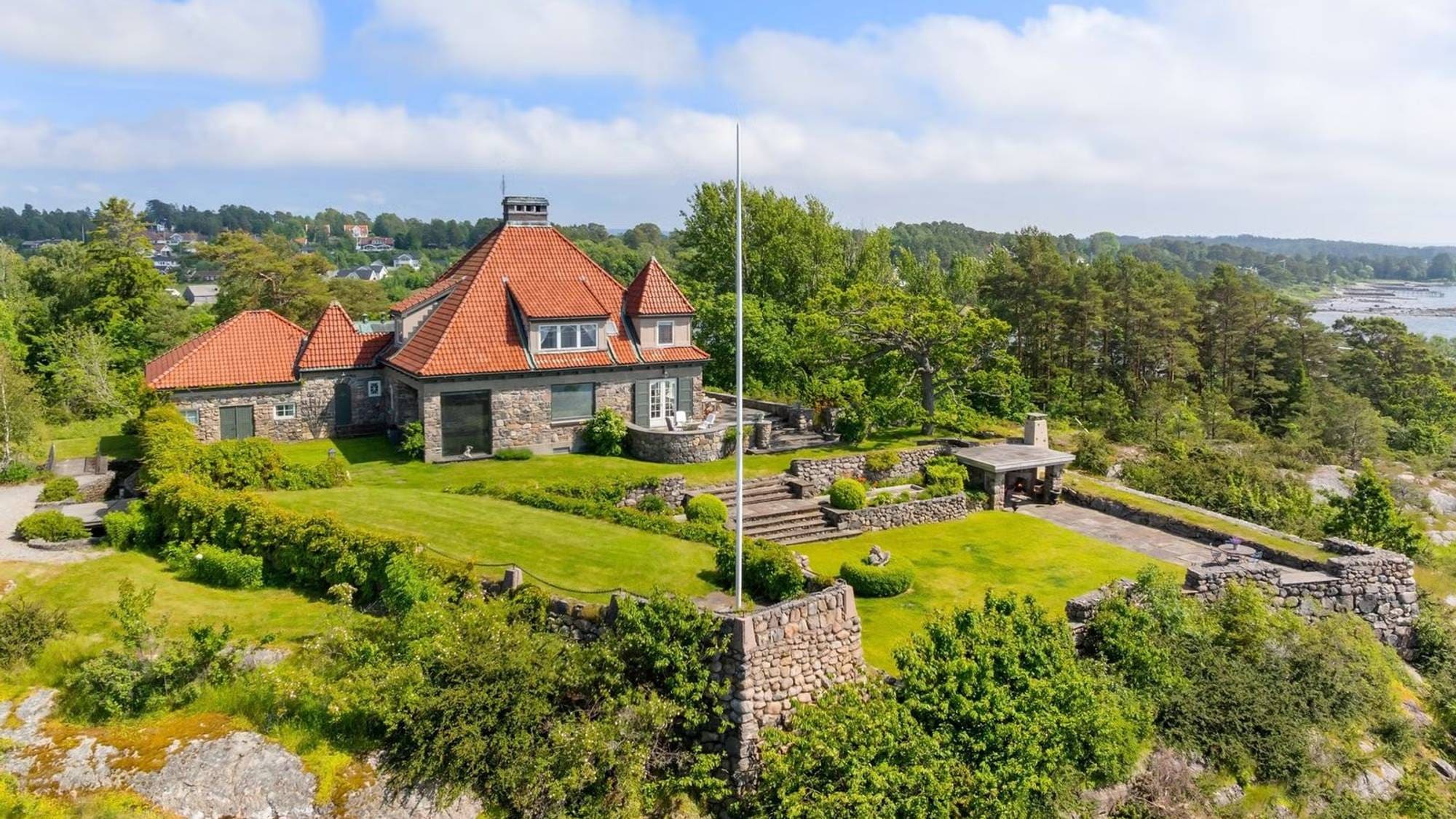 Enebolig i stein med rødt tegltak plassert på en høyde ved sjøen i grønne omgivelser. (Bildebeskrivelsen er laget av en KI-tjeneste)