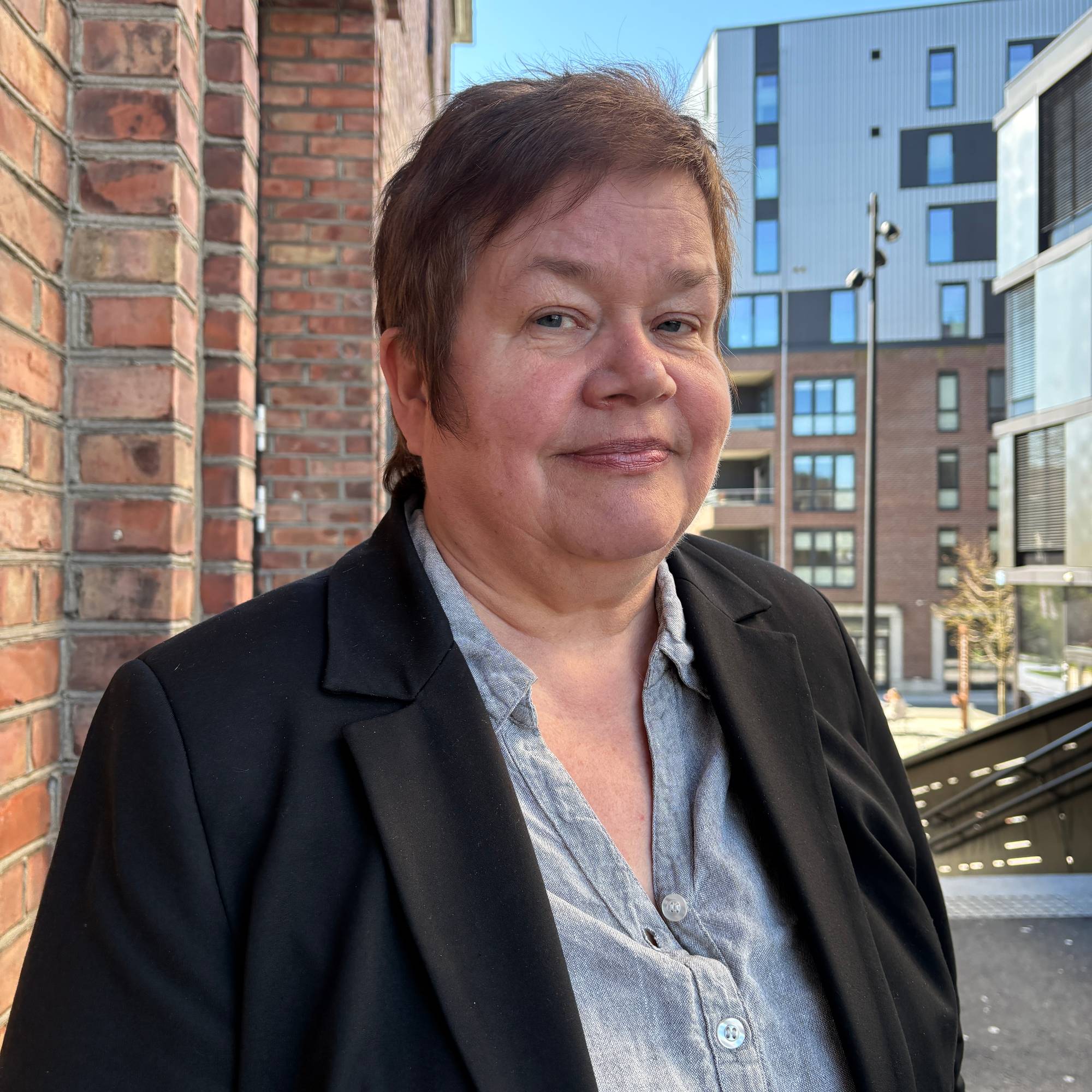 Kvinne med kort brunt hår står foran en teglsteinsvegg med moderne leilighetsbygg i bakgrunnen. (Bildebeskrivelsen er laget av en KI-tjeneste)