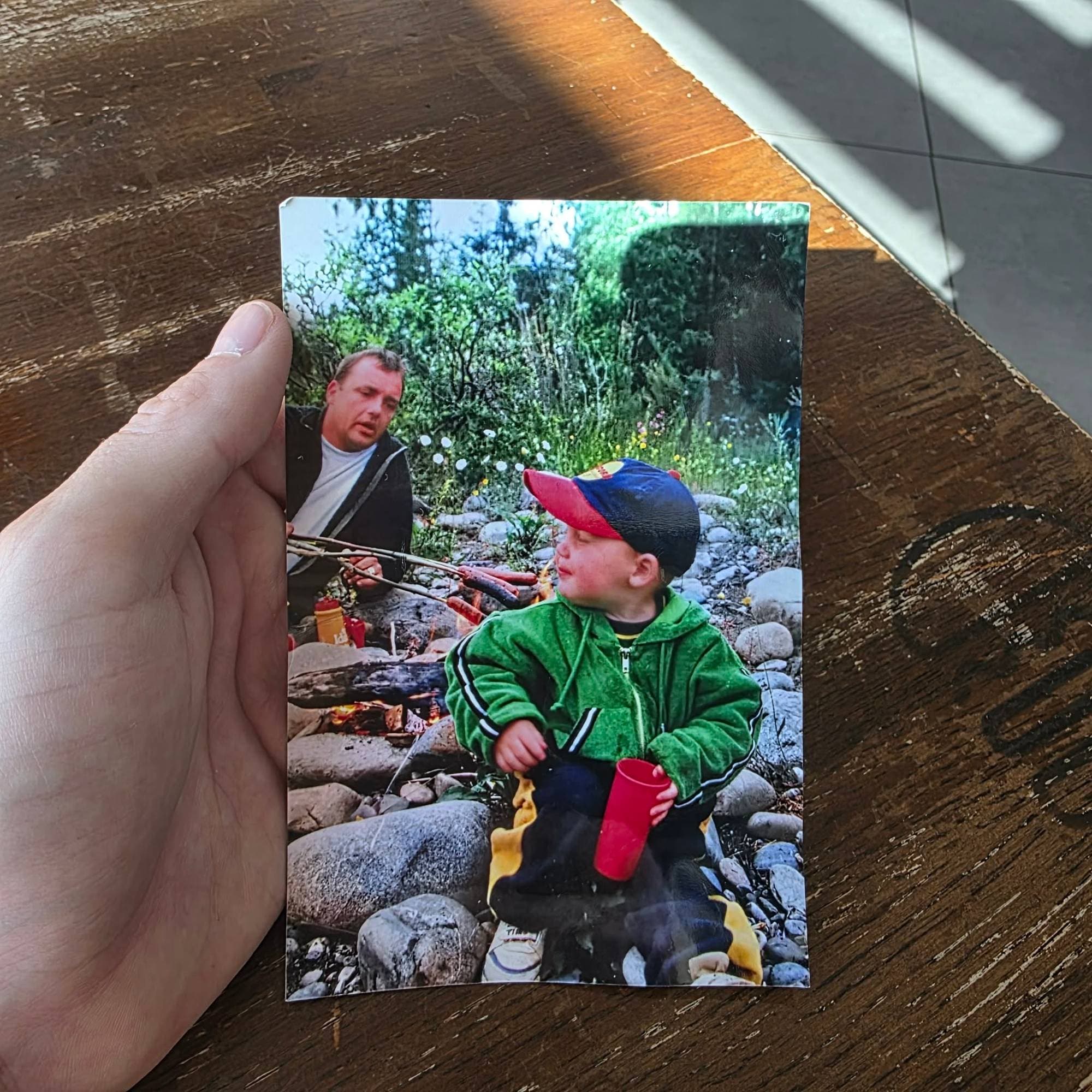 En hånd holder et fotografi av en voksen mann og et barn som griller pølser ved et bål utendørs. (Bildebeskrivelsen er laget av en KI-tjeneste)