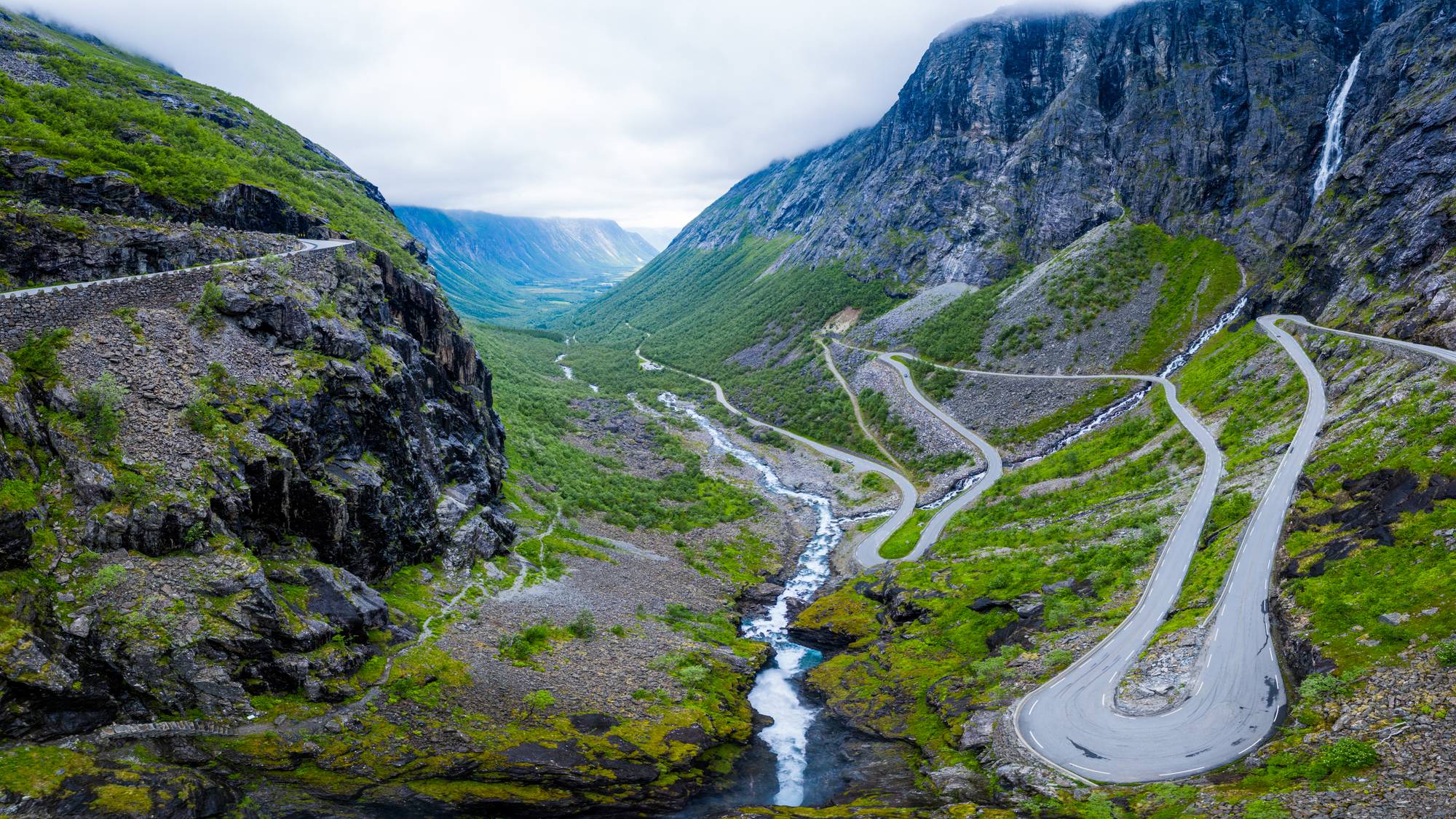 Bildet viser en spektakulær dal med bratte fjellsider og en svingete vei som går langs siden av fjellet. I bunnen av dalen renner en elv, omgitt av grønt vegetasjon og steinete områder. Skyene henger lavt over fjellene, noe som gir et dramatisk inntrykk av landskapet. Veien i bildet er en del av en populær turistattraksjon i området, Trollstigen. (Bildebeskrivelsen er laget av en KI-tjeneste)