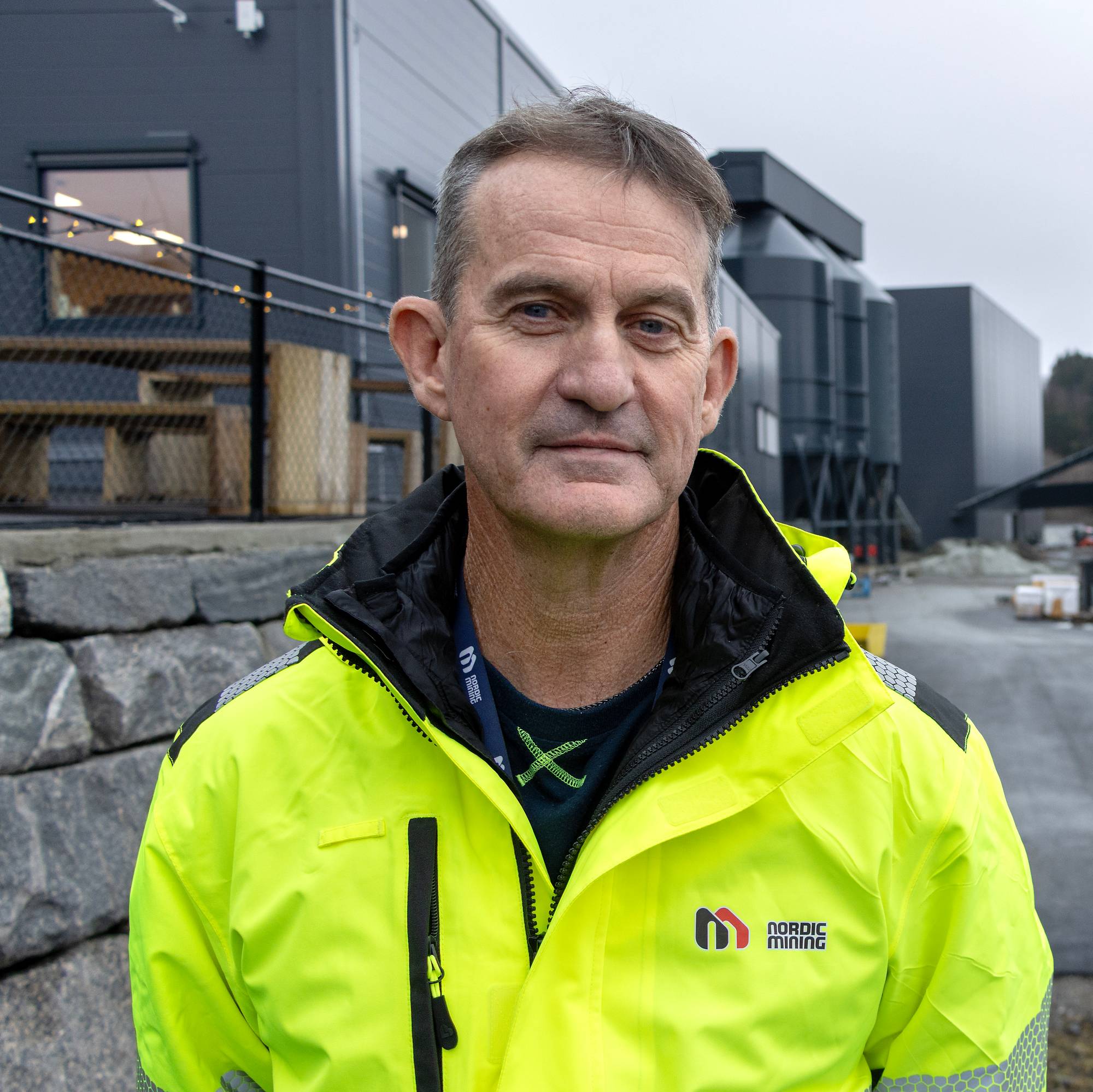 En person iført en lys gul jakke står foran en industriell setting med store, mørke bygninger. Området har en asfaltvei med parkerte biler til høyre og en steinlagt kantstein til venstre. Bakgrunnen viser en fjord og grå himmel, som gir en dyster, men rolig atmosfære. Det er tydelig at bildet er tatt i et industrielt miljø.