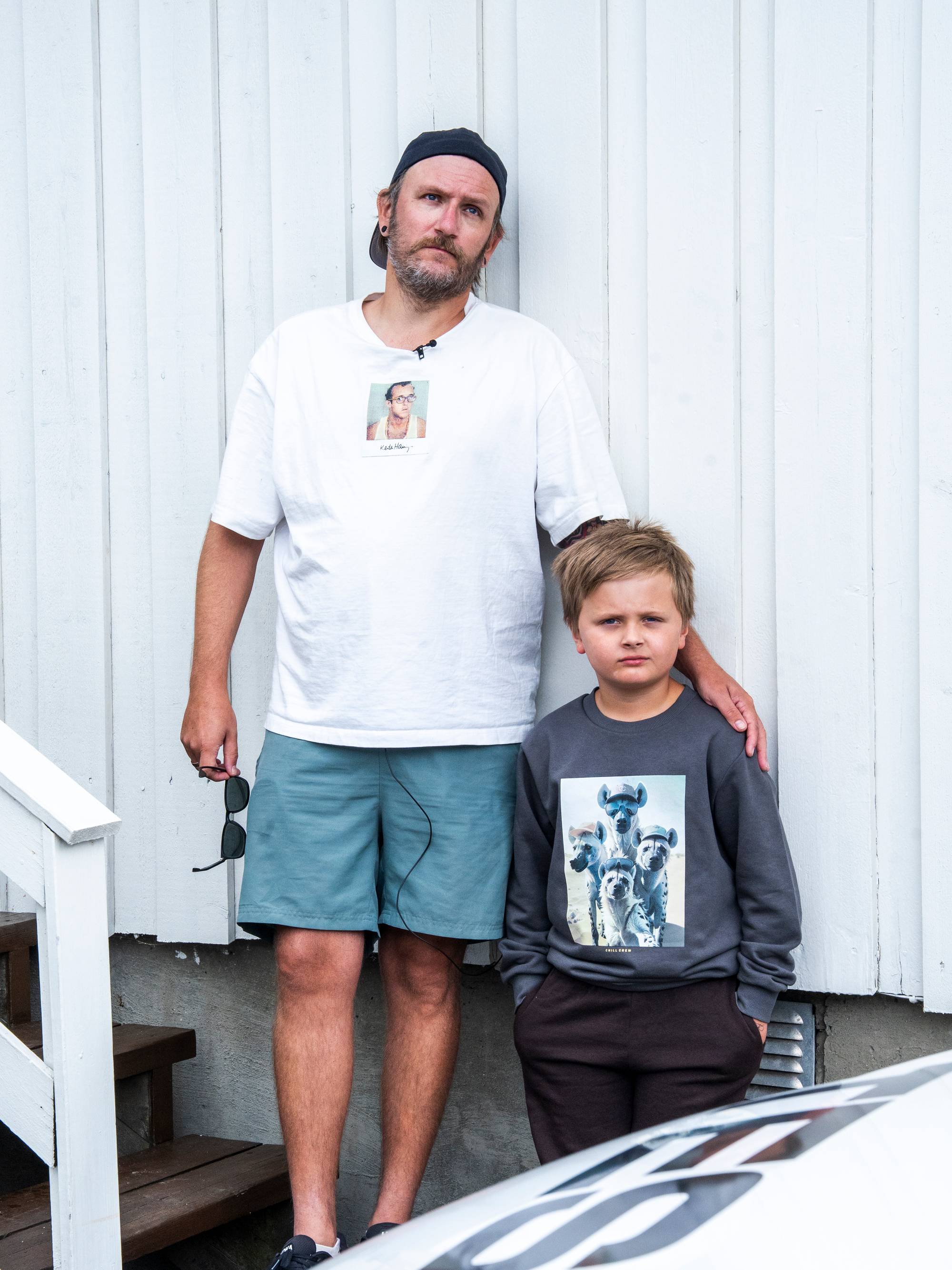 En mann står ved siden av en gutt foran en hvit trevegg. (Bildebeskrivelsen er laget av en KI-tjeneste)