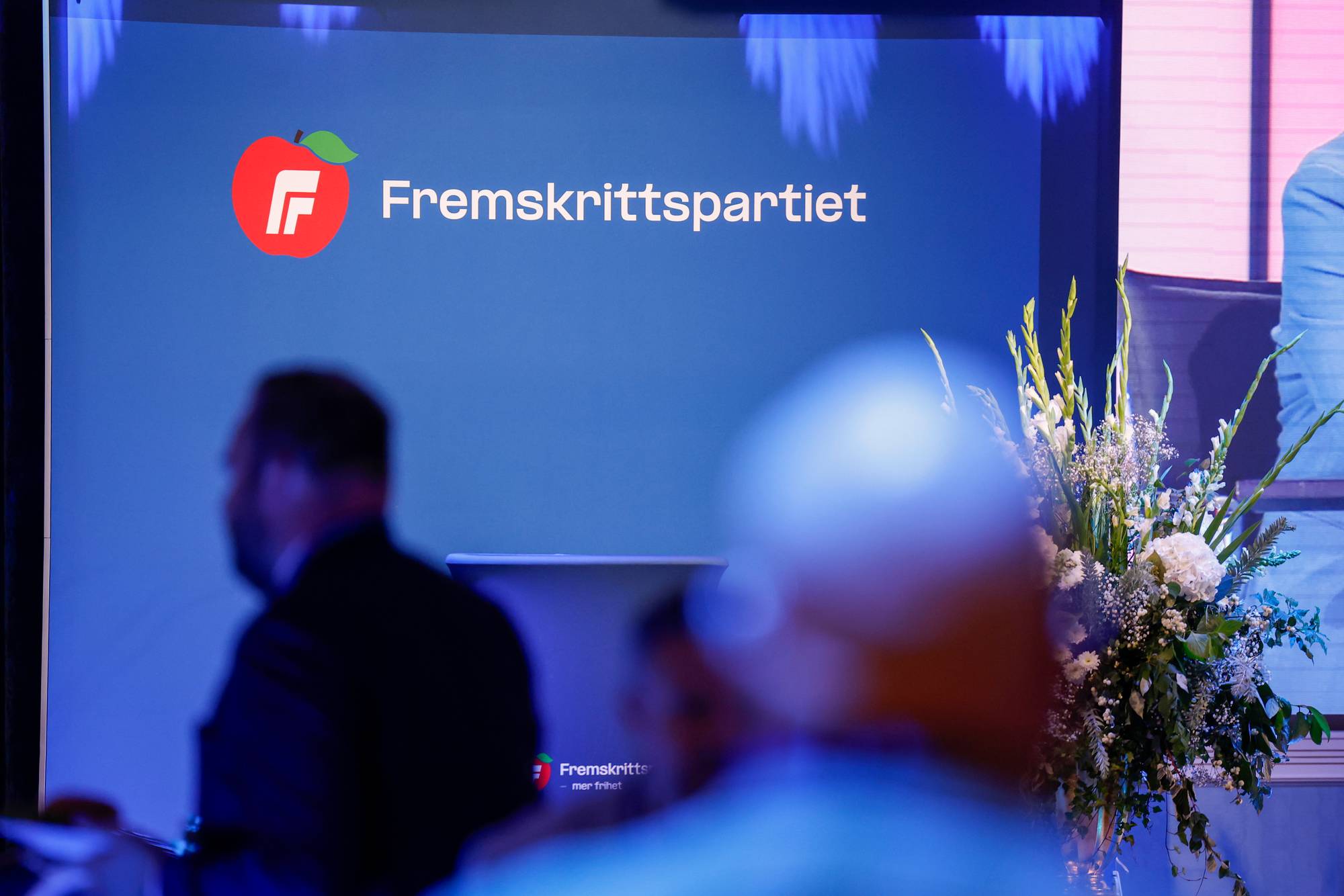En talerstol med logo for Fremskrittspartiet. Deltakere er i forgrunnen. (Bildebeskrivelsen er laget av en KI-tjeneste)